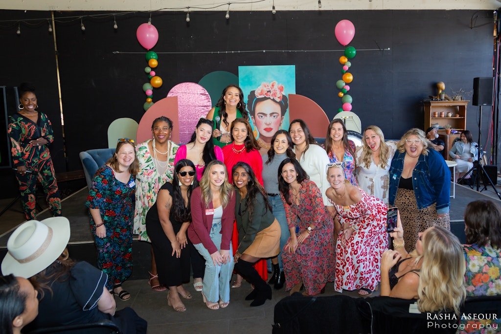 A group of women, some wearing colorful dresses, pose for a group photo at a celebration or event. Pink and green balloons decorate the background, with a Frida Kahlo poster on the wall, and the event appears festive with people taking pictures and s