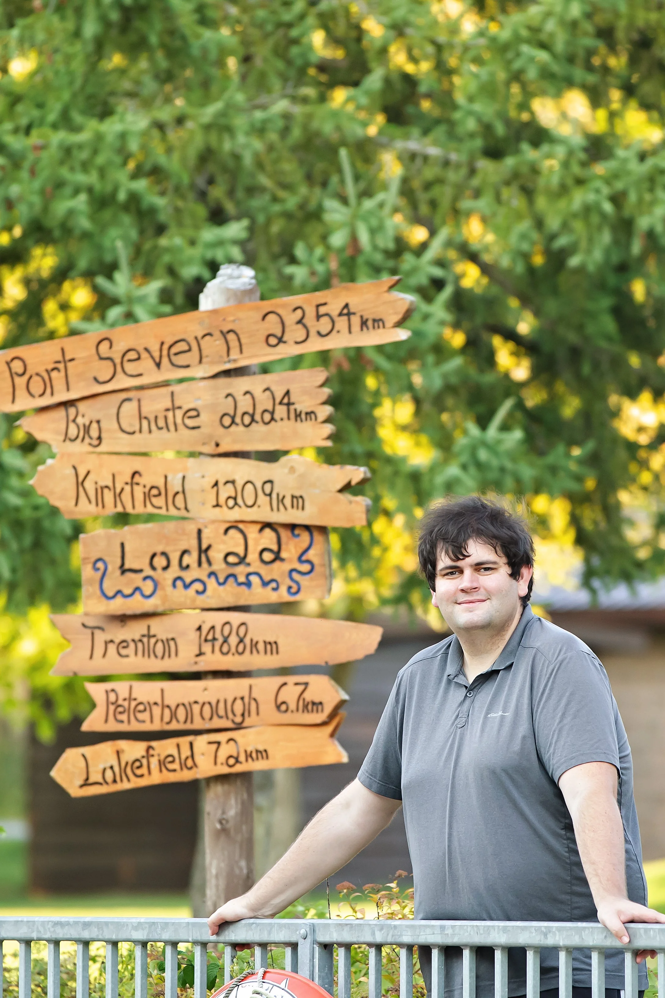 Our Creative Director Jack standing outdoors next to a wooden signpost with multiple directional signs indicating distances to various locations, with trees and a building in the background.