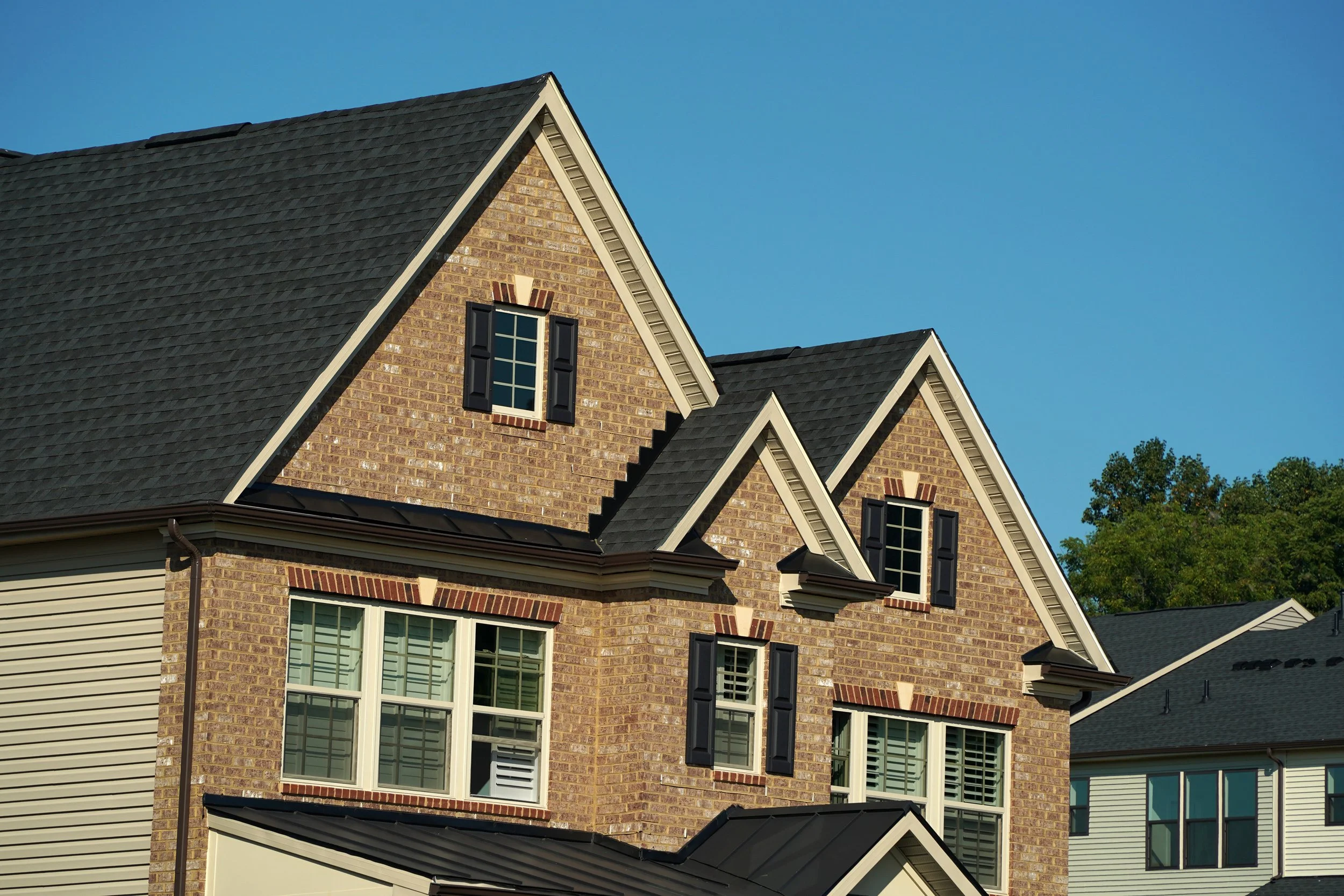 A multi-story house with brick and siding exterior, black-shuttered windows, and a dark gray roof, set against a clear blue sky and some green trees.