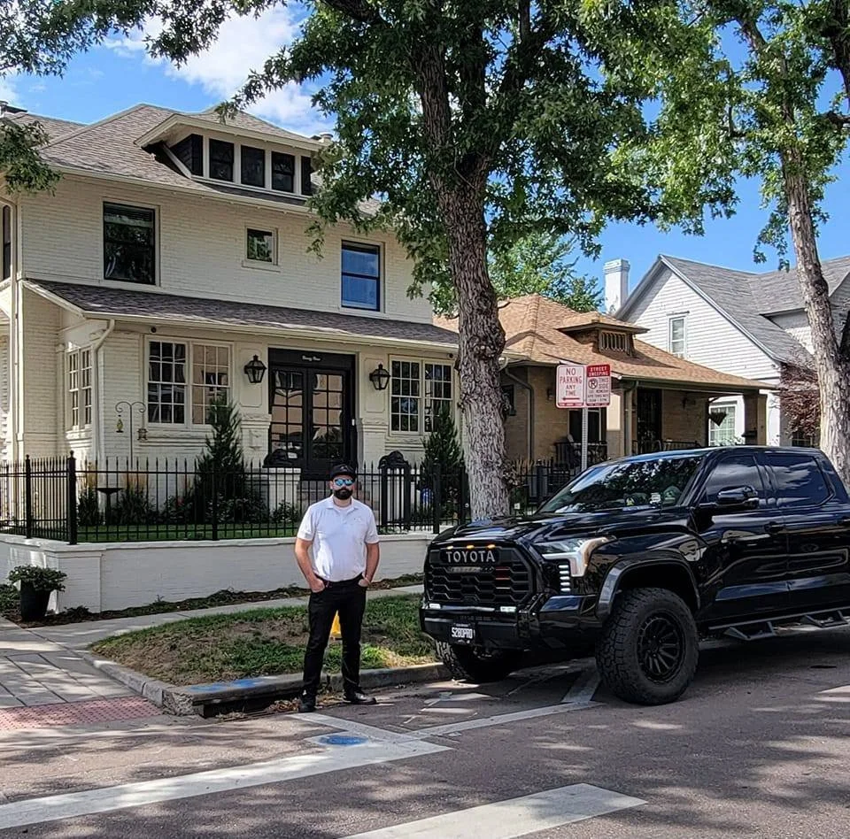 A man standing on the sidewalk near a black Toyota truck in front of a residential house with a large tree in the yard. The man is wearing a white shirt, black pants, and a face mask. There are parking regulation signs on a pole behind him.