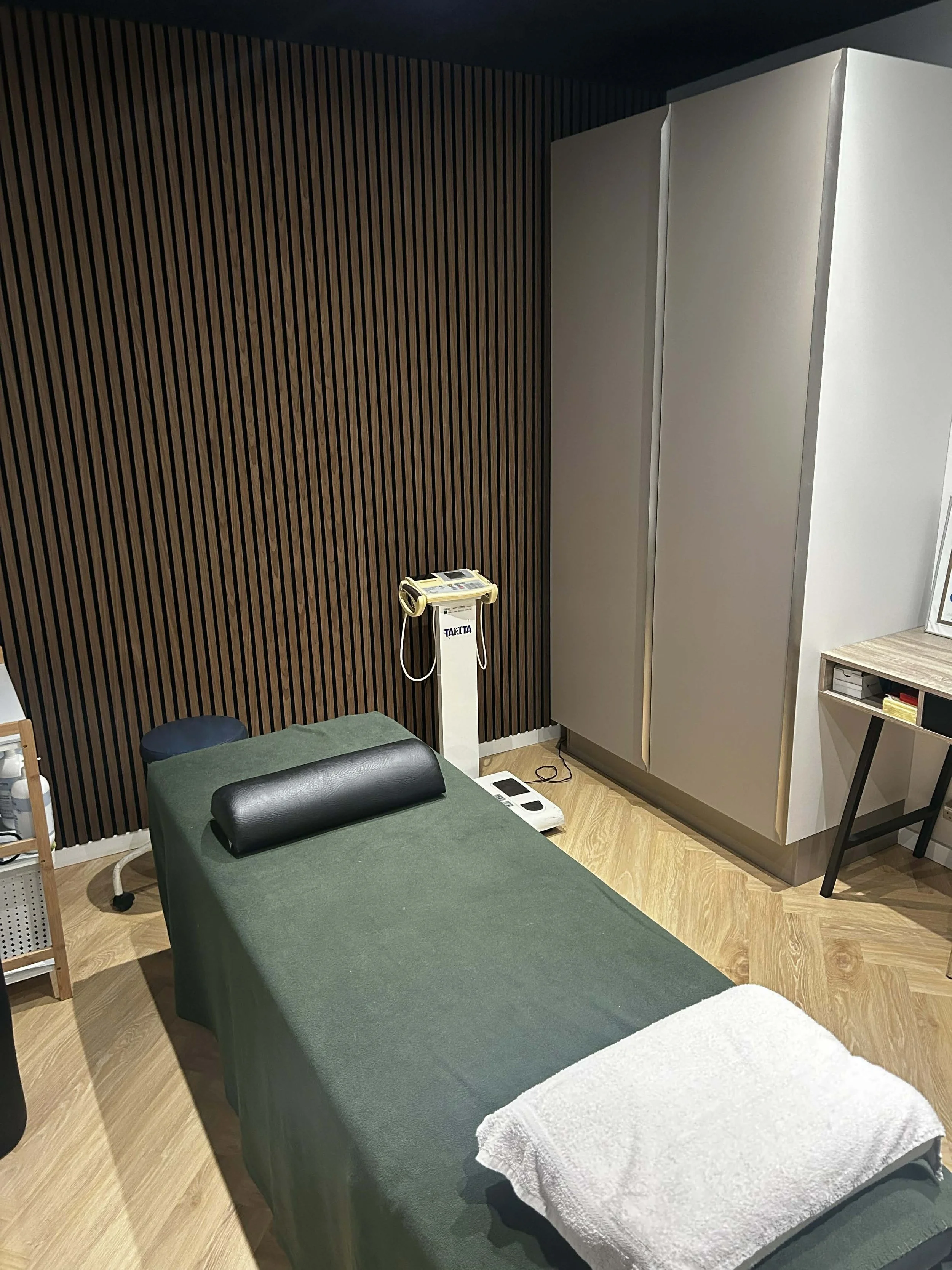 Massage table with a black pillow, a white towel at the end, a chair, a small storage cart, a scale, and a wooden floor in a therapy room with dark wood panel wall and white cabinet.