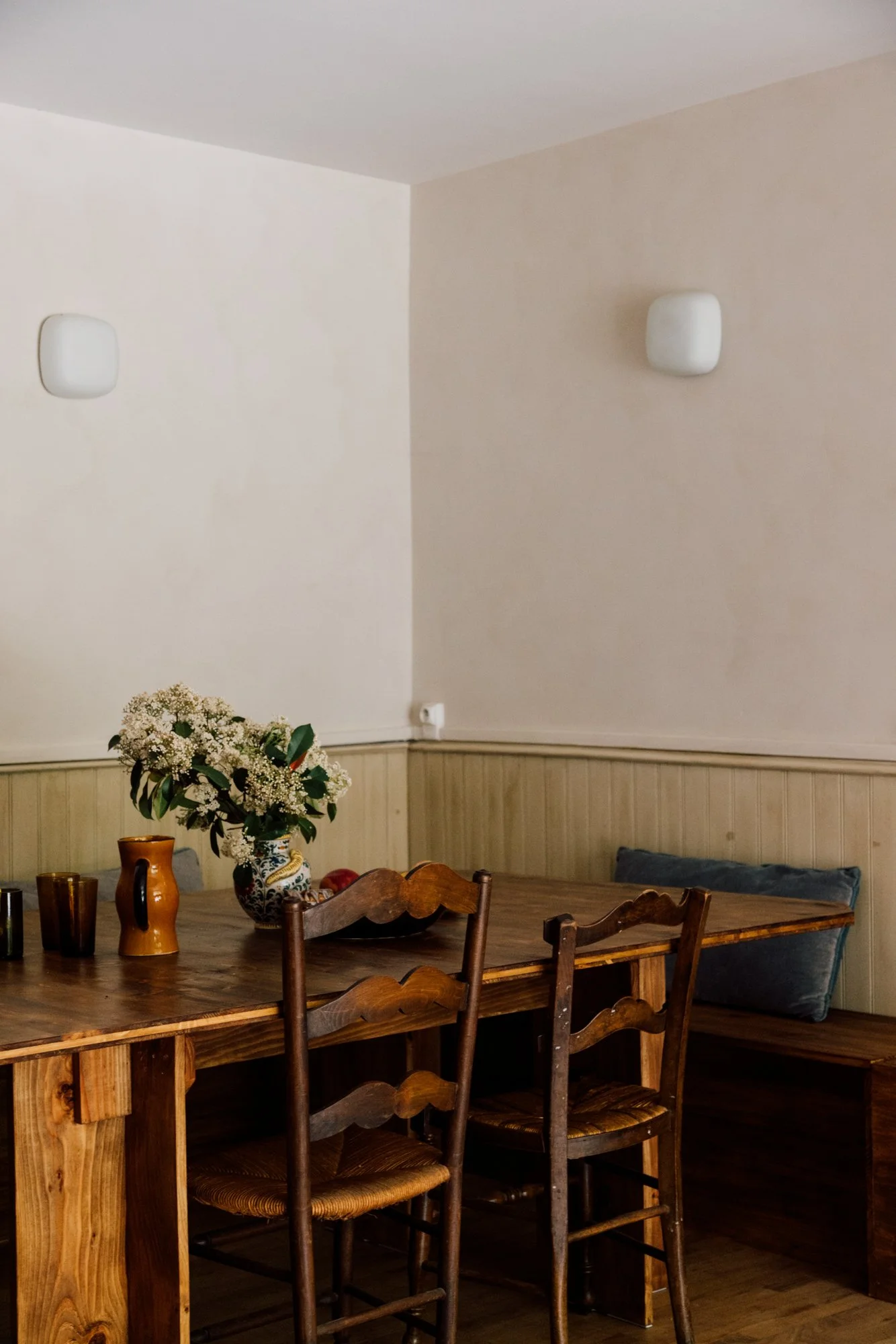 Une pièce avec une table en bois autour de laquelle se trouvent plusieurs chaises en bois. Sur la table, il y a un vase avec des fleurs blanches et quelques verres et objets en céramique. Le mur en arrière-plan est partiellement en bois et peint en b