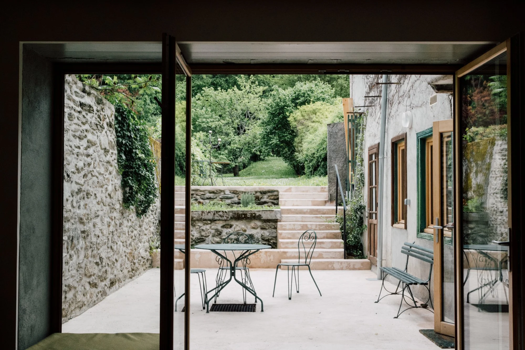 Vue d'une terrasse extérieure avec mobilier en métal, escaliers menant à un jardin verdoyant, murs en pierre et fenêtre avec cadre en bois.