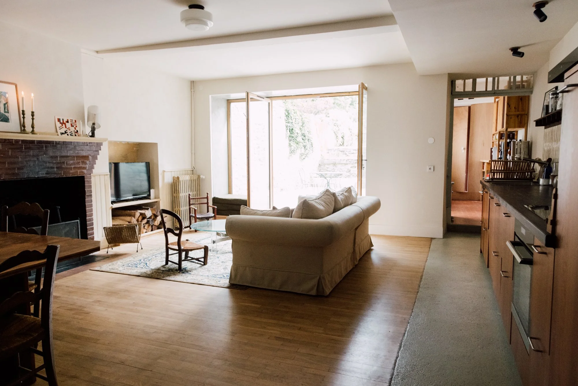 Salon intérieur avec cheminée, télévision, canapé beige, petits fauteuils, grande baie vitrée laissant entrer la lumière naturelle, cuisine en arrière-plan.