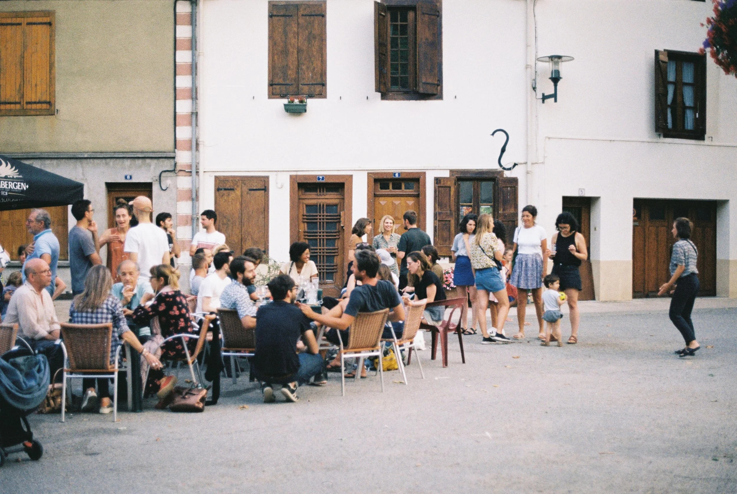Une scène de rassemblement urbain avec plusieurs personnes assises à des tables en plein air, discutant et socialisant, tandis que d'autres personnes se tiennent debout ou marchent dans la rue, devant un bâtiment avec fenêtres à volets en bois.