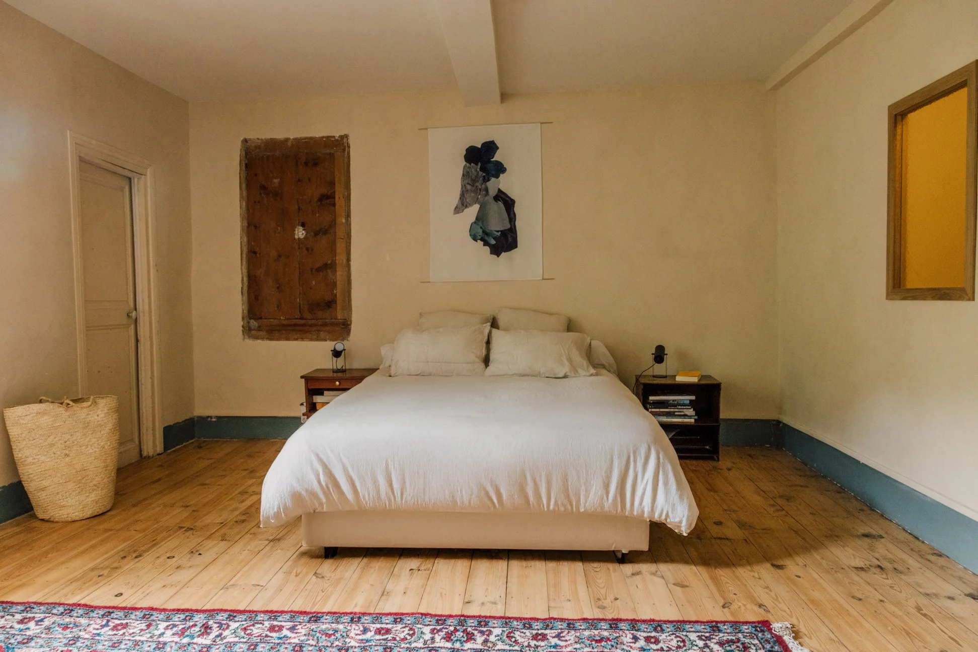 Chambre avec lit blanc, deux tables de nuit avec lampes, tableau abstrait au mur, porte ancienne, tapis au sol