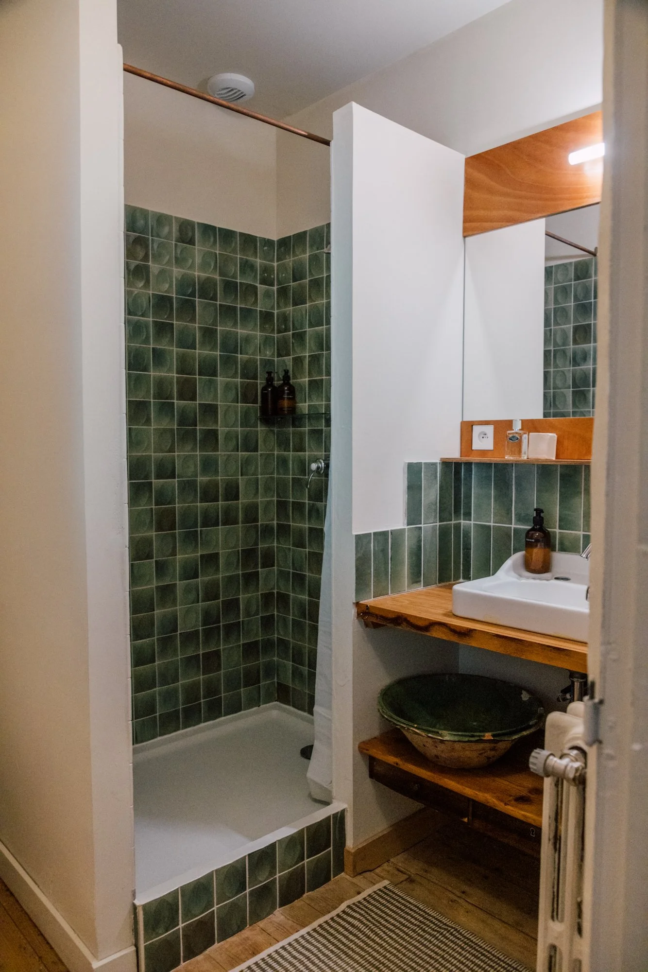 Petite salle de bain avec douche en carreaux verts, lavabo blanc sur un meuble en bois, miroir avec lampes, et éléments en bois décoratifs.