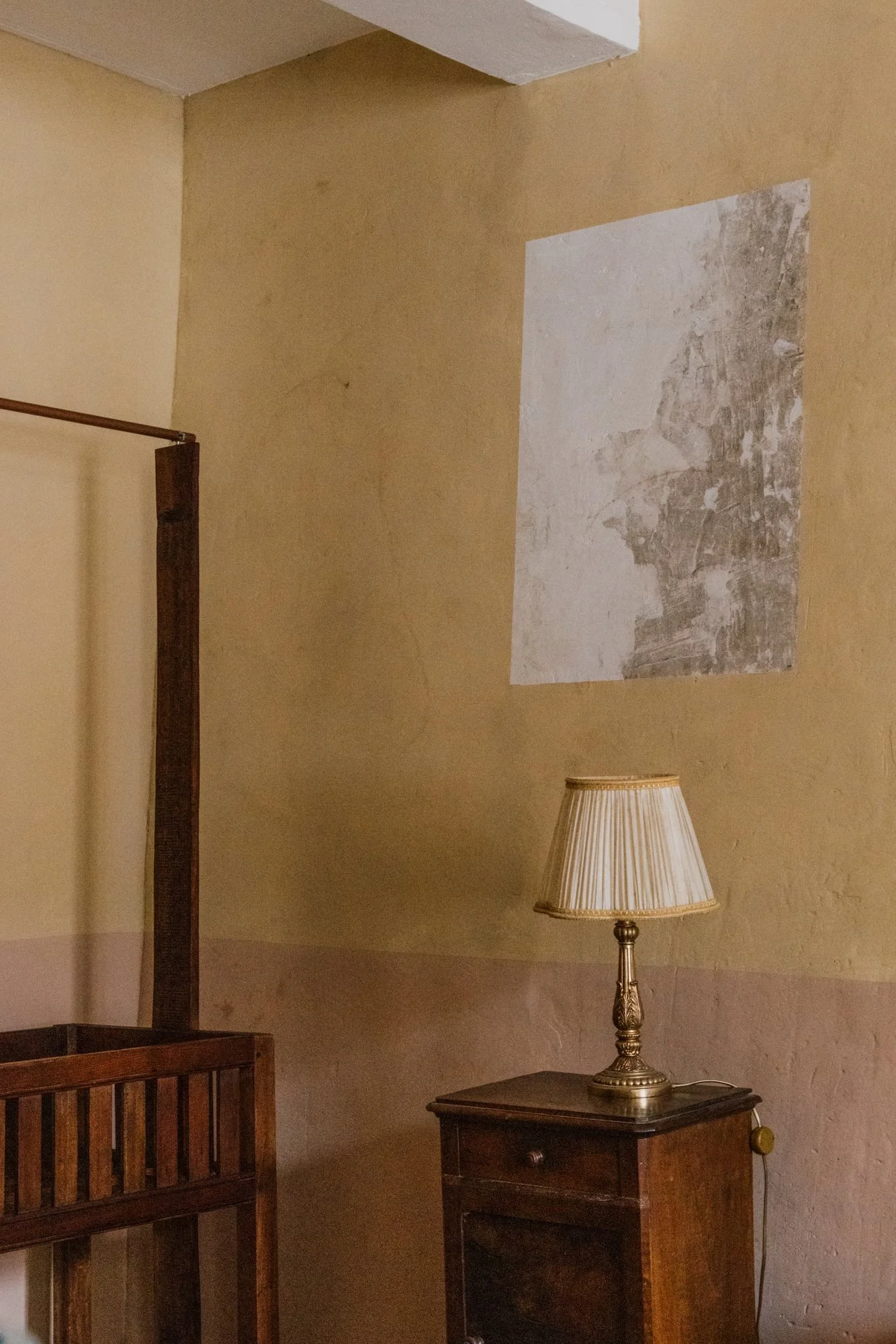 Une lampe de table posée sur une petite commode en bois, dans une pièce avec un mur en face qui a un ravalement partiel de peinture ou de plâtre.