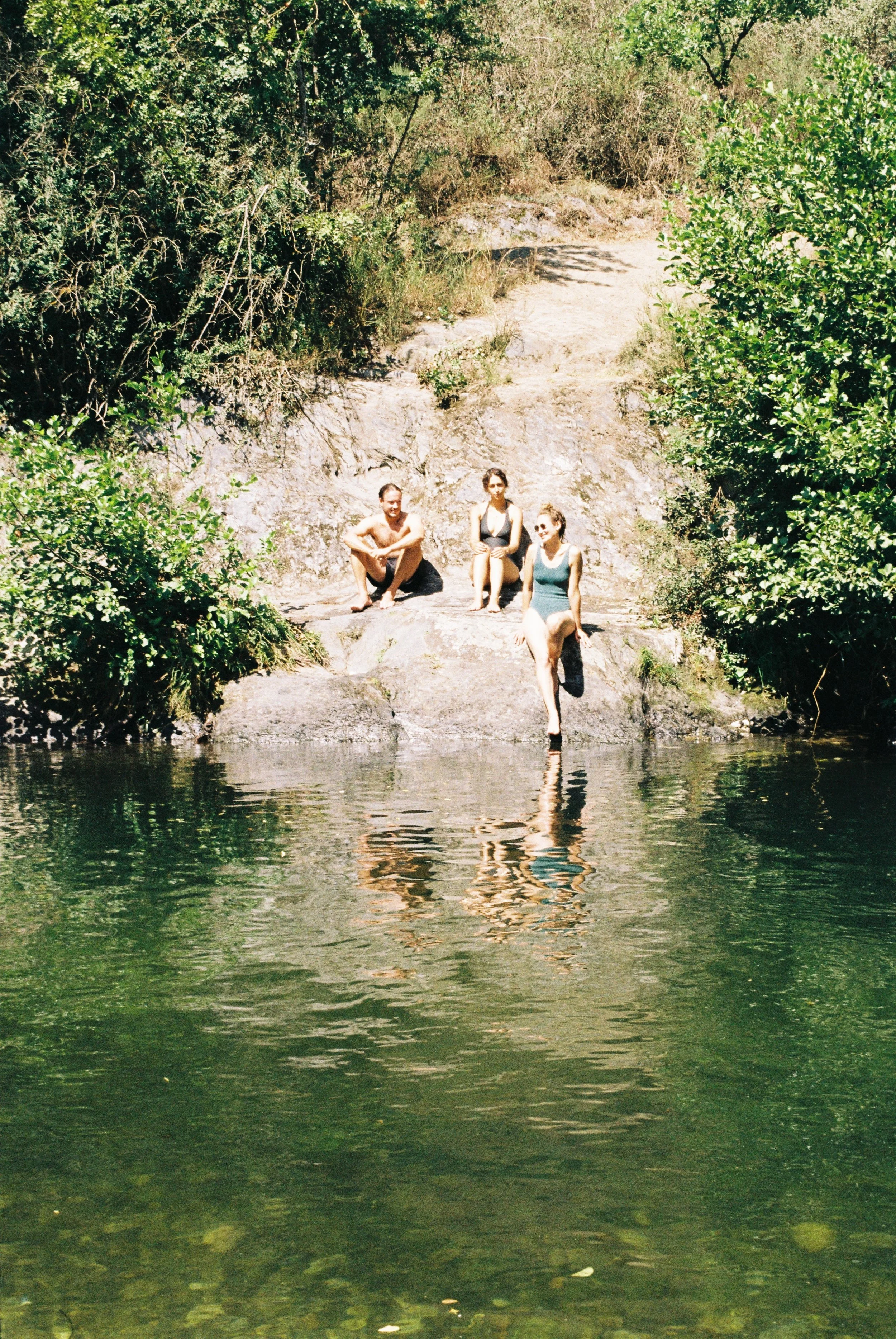 Trois personnes assises sur un rocher près d'une rivière entourée de végétation dense.