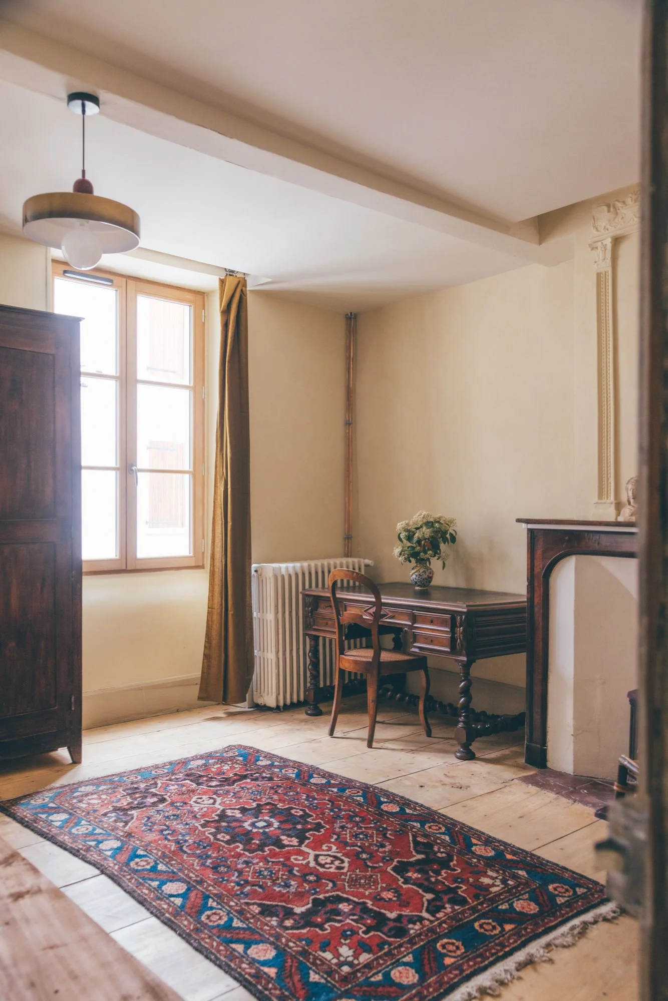 une chambre avec un tapis persan, un bureau en bois, une chaise, une armoire en bois, une fenêtre avec des rideaux dorés, un radiateur et une lampe suspendue.