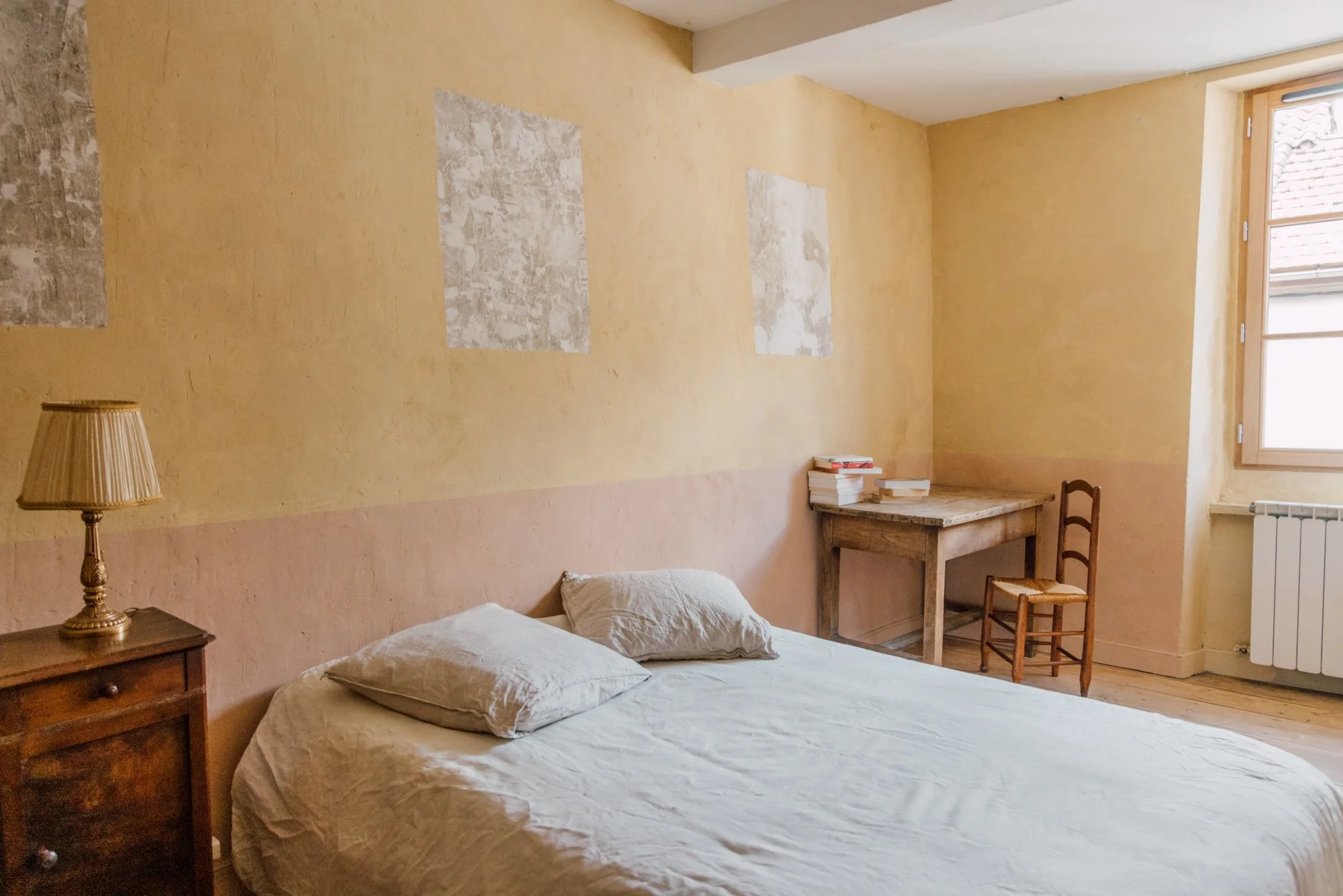 Chambre simple avec un lit, une table de nuit avec une lampe, une fenêtre et un bureau en bois avec une chaise.