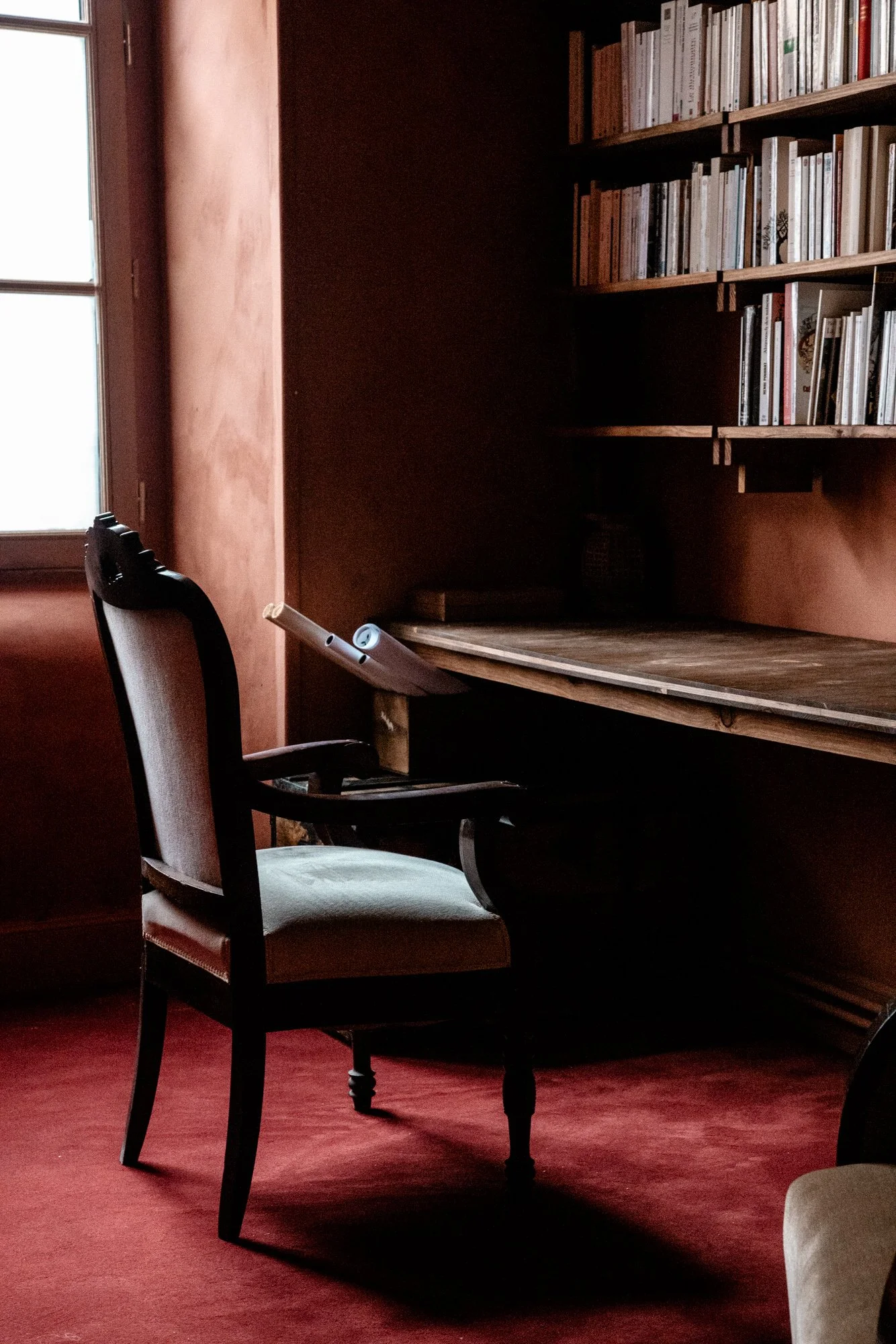 Un bureau en bois avec une chaise ancienne devant une étagère remplie de livres, une fenêtre à gauche laissant entrer la lumière.