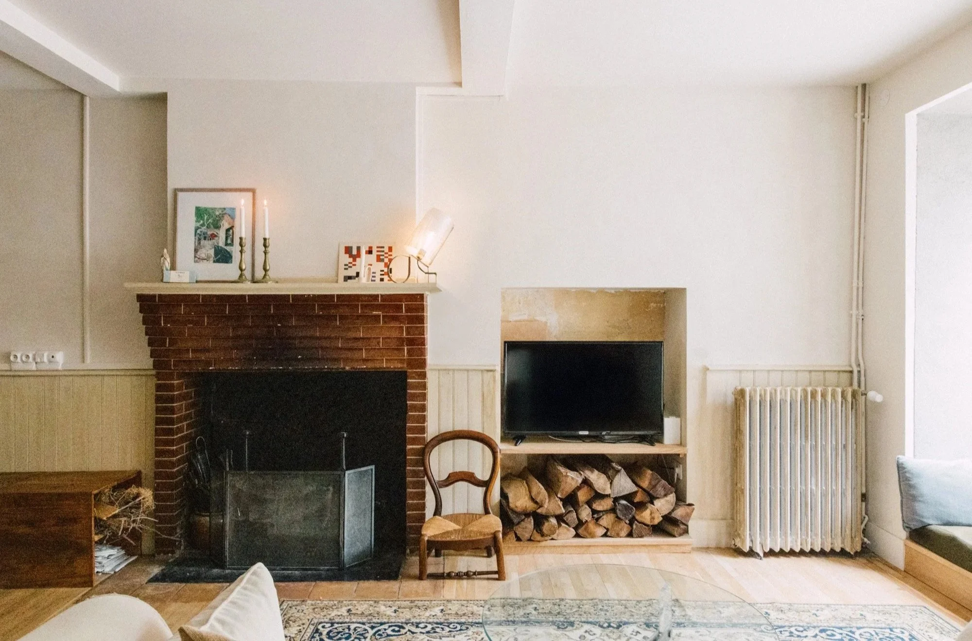 Salle de séjour avec cheminée en briques, un meuble avec du bois empilé, un téléviseur, une chaise en bois, un radiateur, et divers objets décoratifs sur la cheminée.