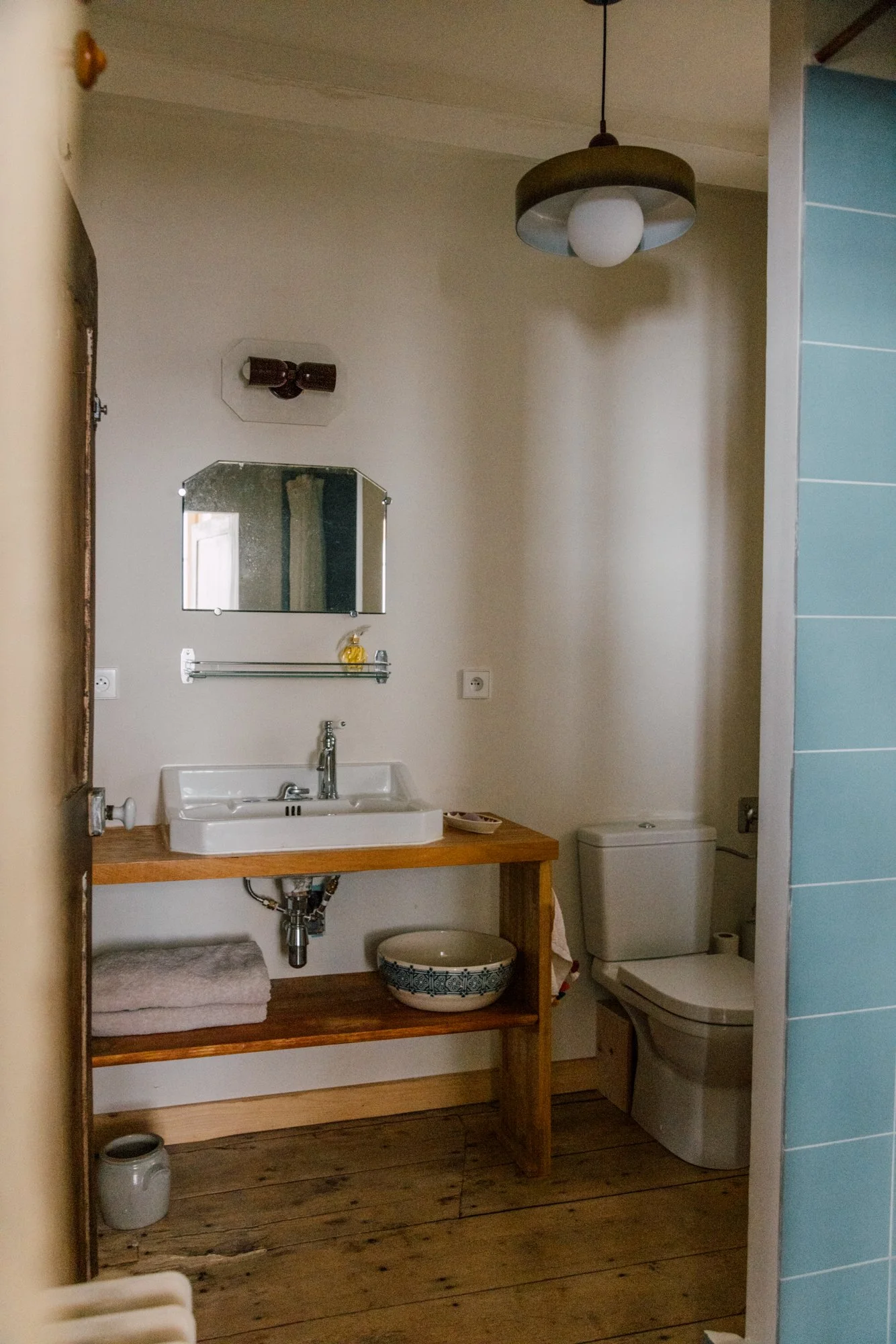 Une salle de bain simple avec un lavabo en céramique blanc placé sur un meuble en bois, un miroir au mur, un toilette à côté, une étagère avec des serviettes et un bol décoratif, un plafond avec un luminaire suspendu