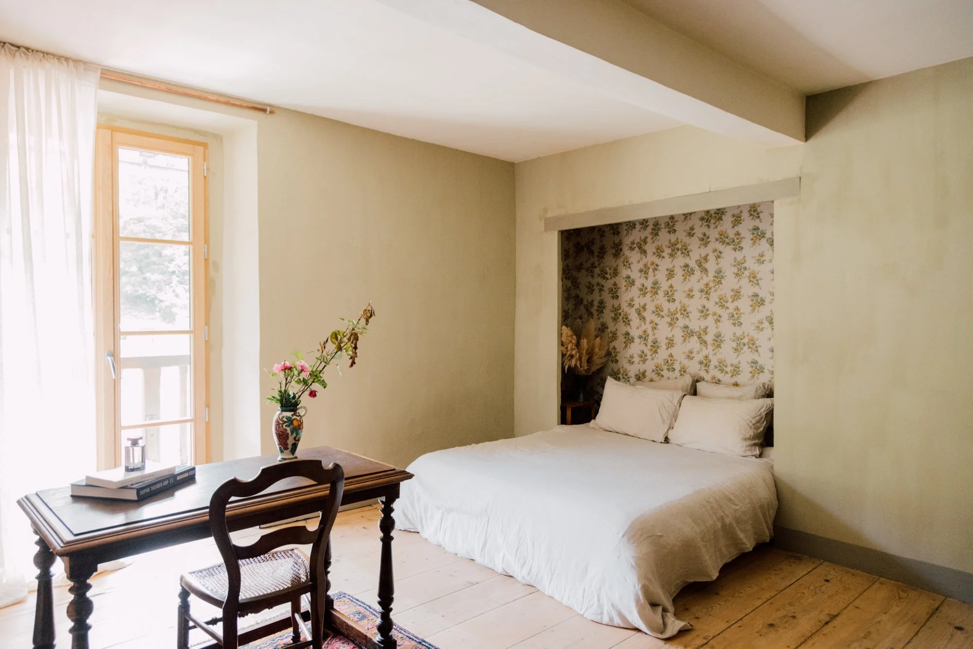 Chambre lumineuse avec lit blanc, décor floral derrière la tête de lit, table en bois avec vase de fleurs, fenêtre laissant entrer la lumière