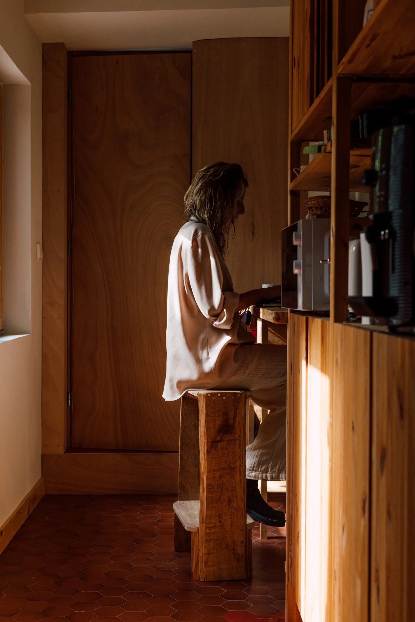 Une femme s'assoit sur un banc en bois dans une cuisine en bois, regardant un téléphone, avec une lumière naturelle tombant de côté.