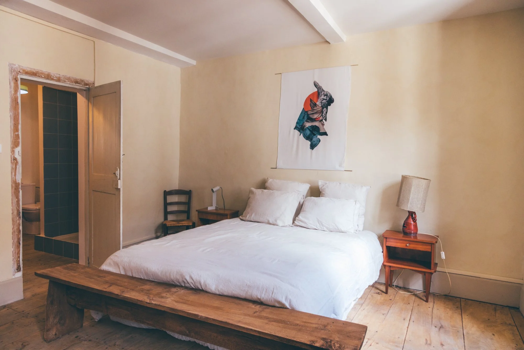 Chambre avec lit double, deux oreillers, une table de chevet avec lampe, un tableau décoratif sur le mur, lampe sur la table de chevet et une chaise en bois. Partie d'une salle de bain avec toilettes visible à travers une porte ouverte.