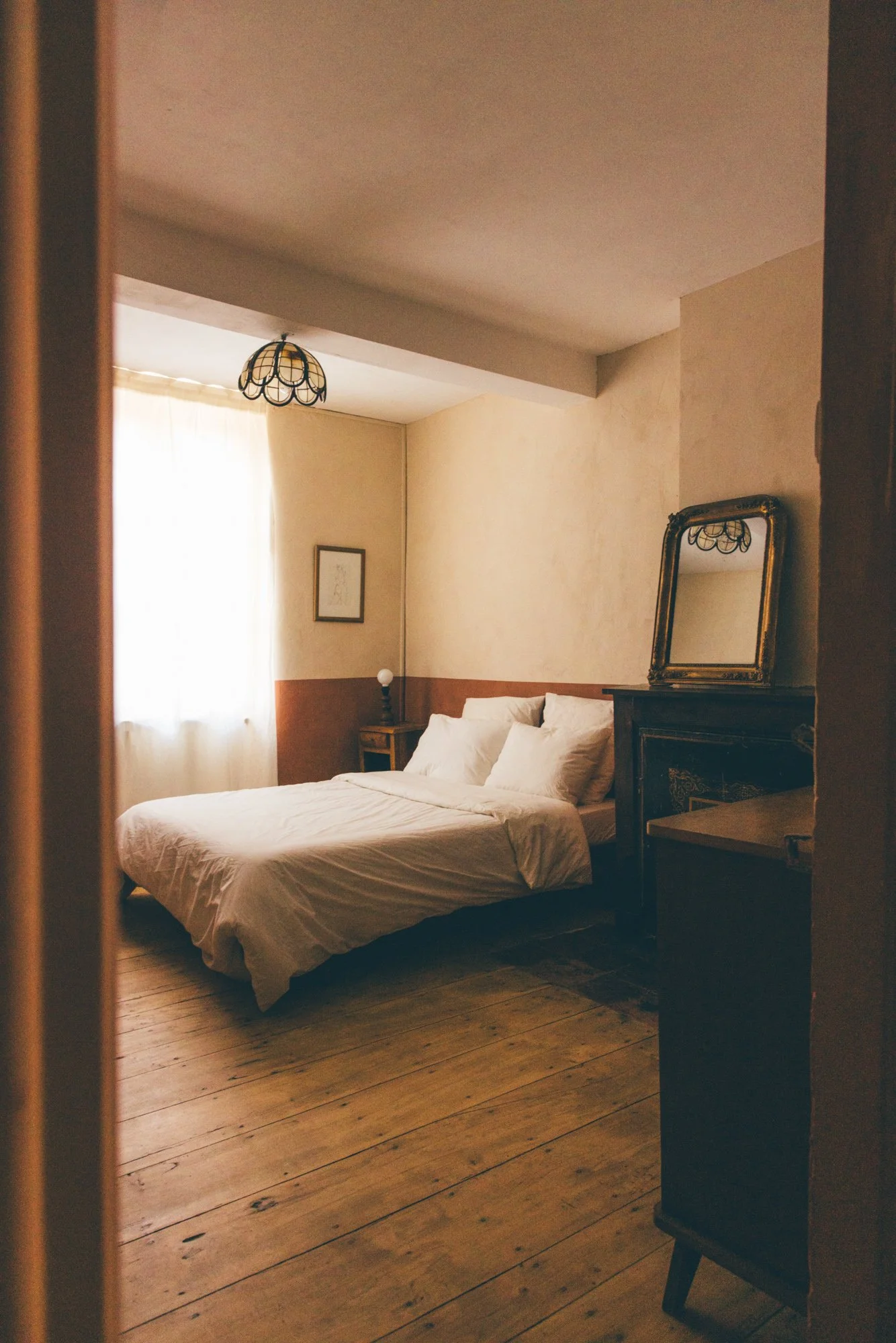 Chambre avec un lit blanc, une fenêtre avec rideaux, un miroir ancien et un éclairage tamisé.