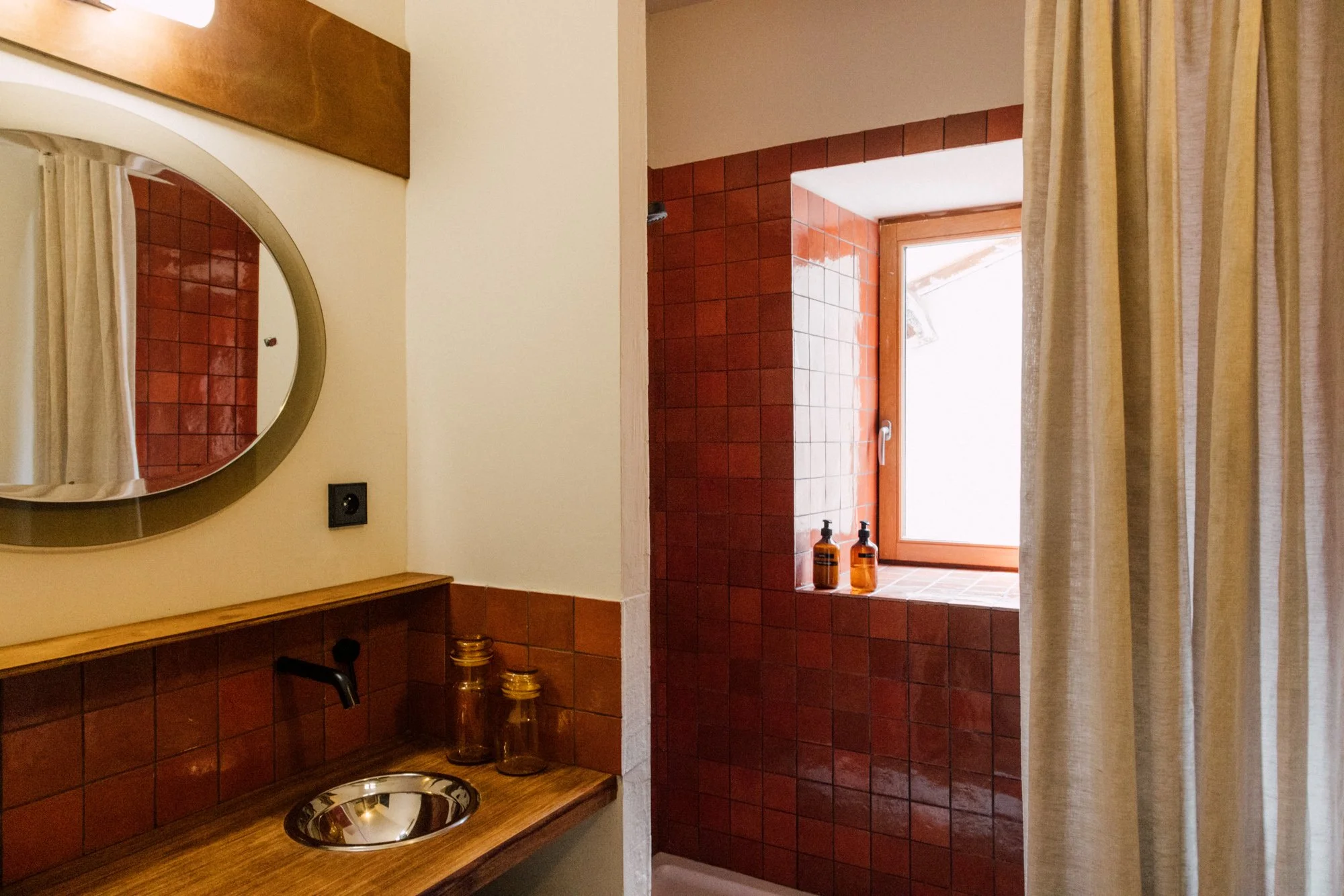 Espace douche avec fenêtre en bois, murs en carrelage rouge et accessoires de bain en verre à côté d'un miroir rond.