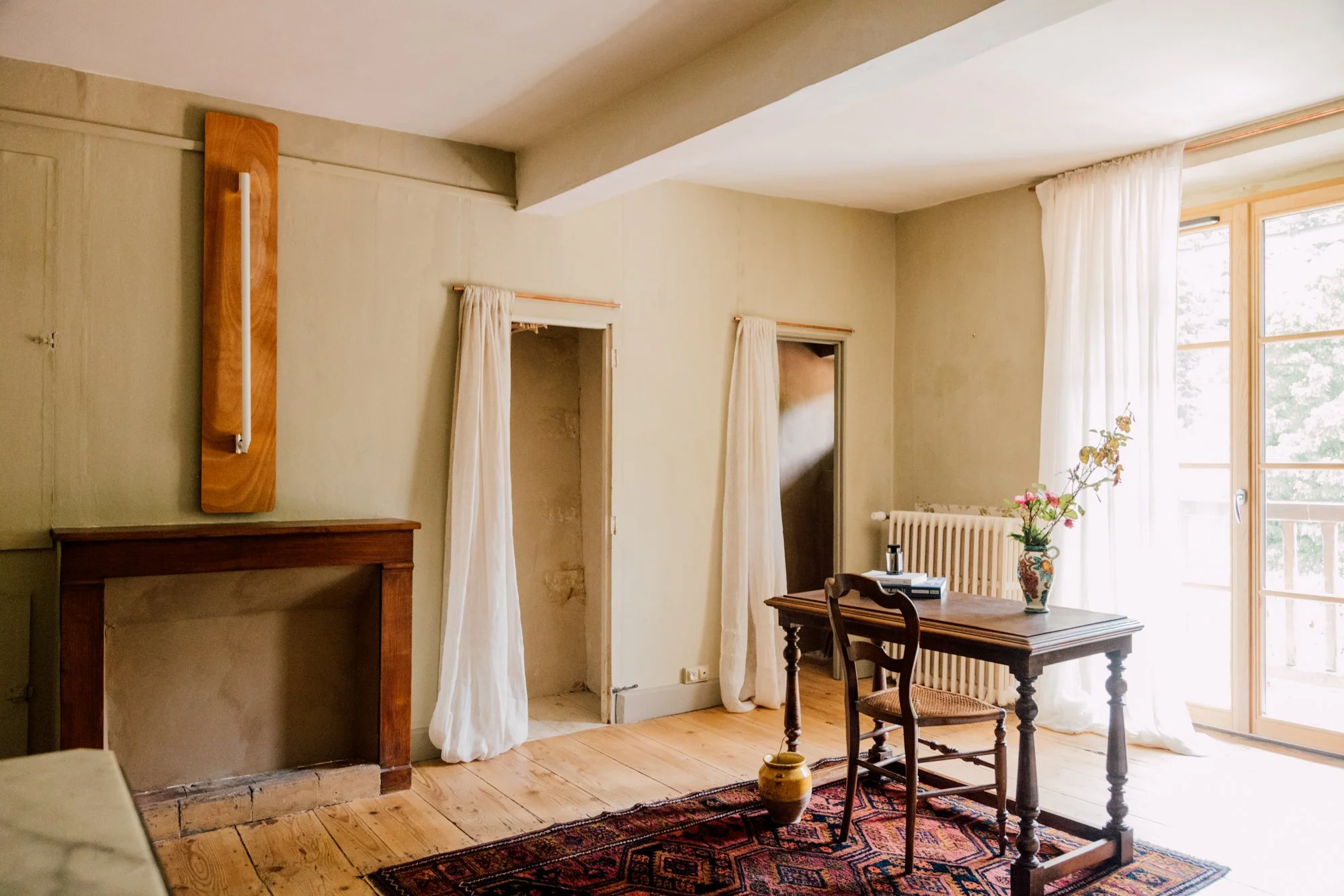 Salon avec cheminée, table en bois, deux chaises, vase avec fleurs, fenêtre avec rideaux blancs, tapis coloré, porte avec rideaux, et un mur beige
