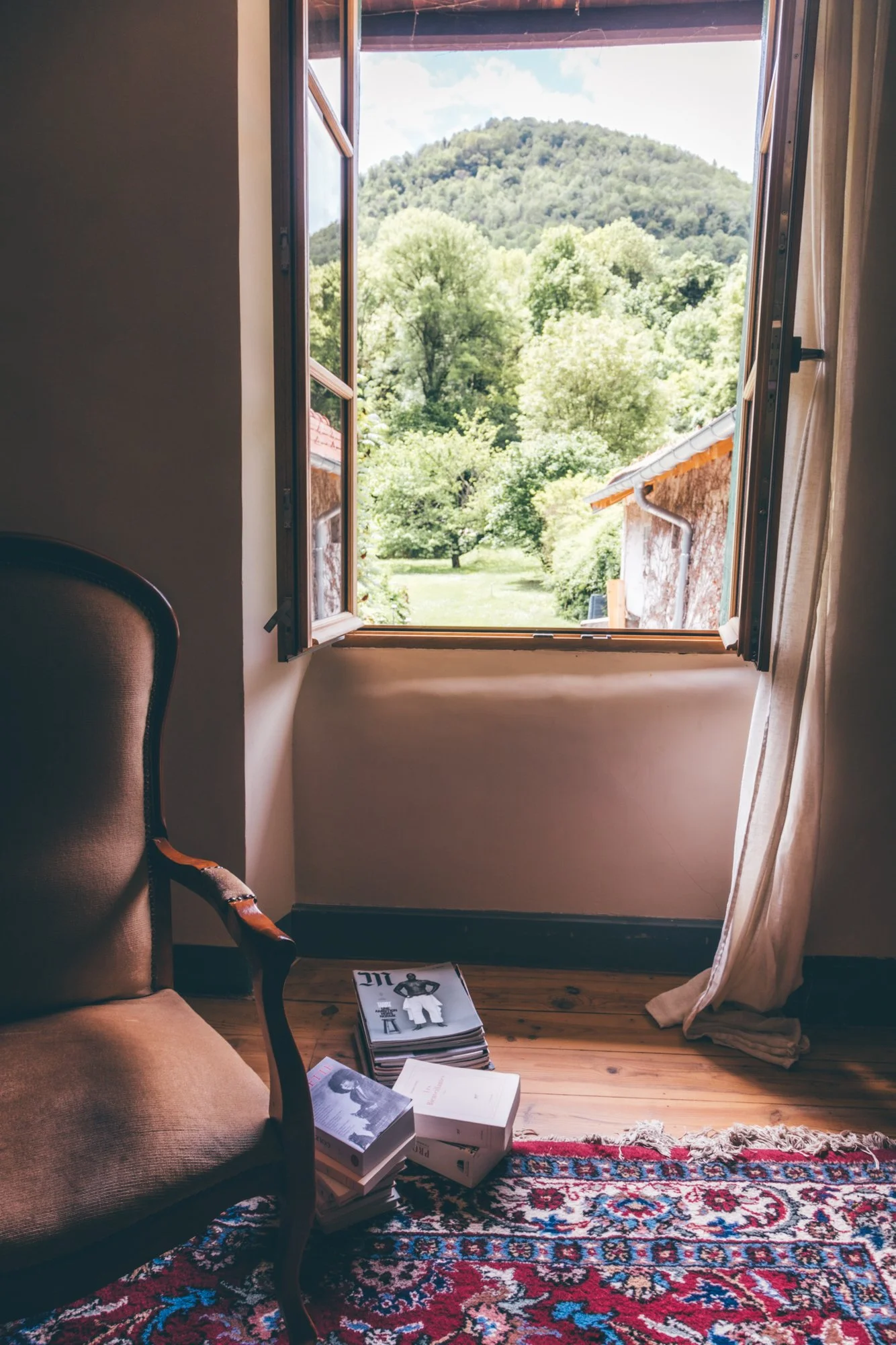 Une chambre avec un fauteuil en bois avec rembourrage beige à gauche, un tapis rouge avec motifs complexes au sol, des livres empilés à côté d'un livre blanc, un mur blanc, et une fenêtre ouverte donnant sur un paysage de collines verdoyantes sous un ciel nuageux.