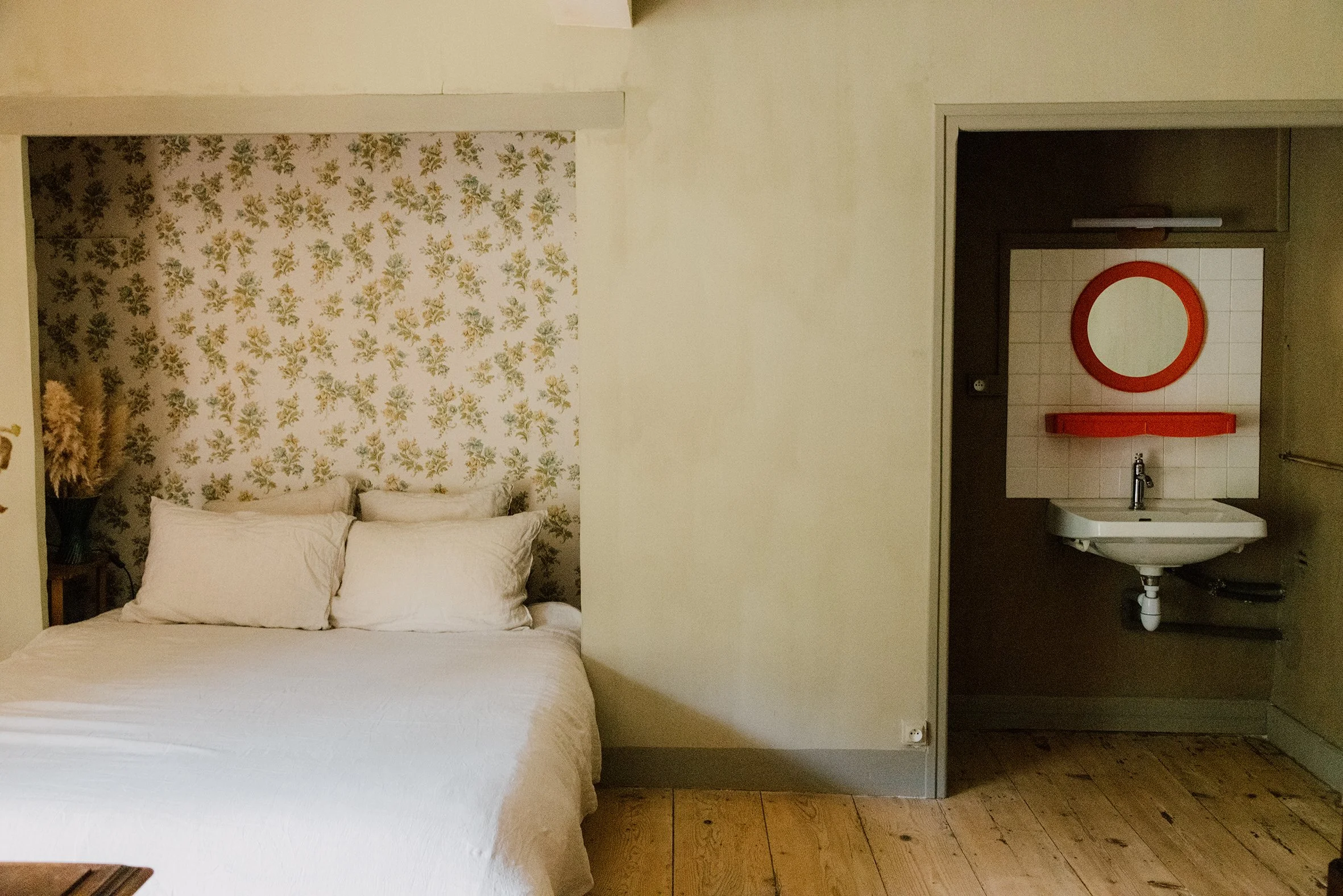 Chambre avec lit blanc et décor floral, mur en papier peint avec motif vert et beige, coin lavabo avec miroir rond rouge et étagère rouge.