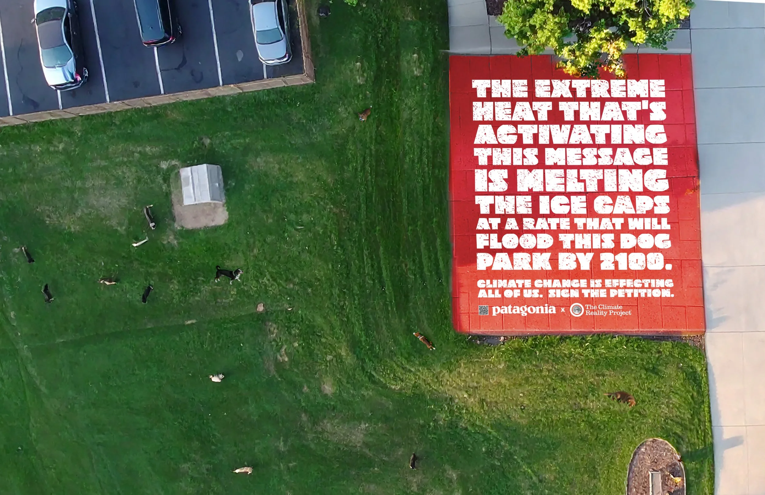 An aerial view of a grassy area with a few small dogs walking around. On the right side, there is a large red wall with white text promoting climate change awareness, stating that extreme heat is melting the ice caps at a rate that will flood a local dog park by 2100. There are parked cars and a sidewalk adjacent to the grassy area.
