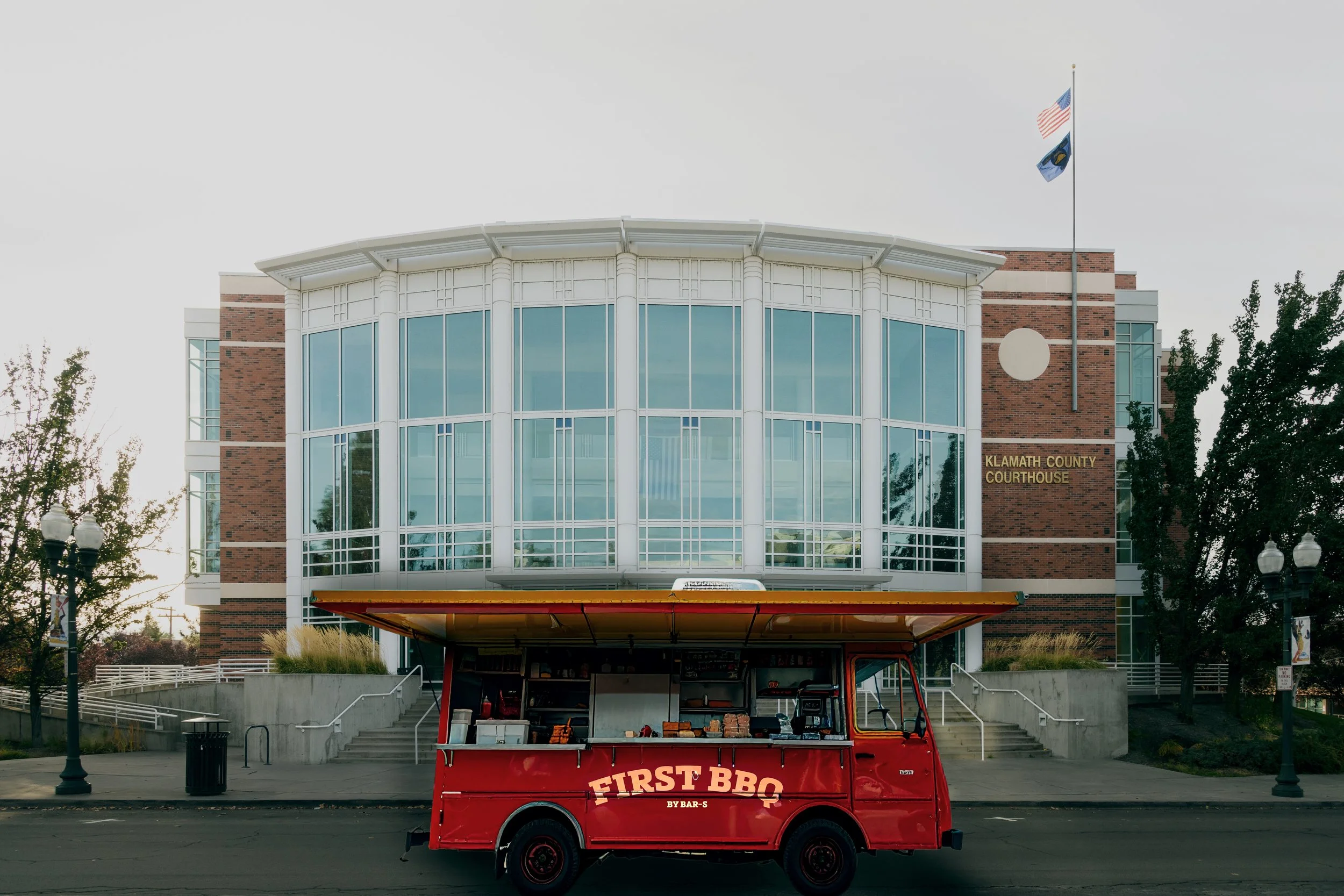 A red food truck labeled 'First BBQ by Bar-S' parked in front of the Klamath County Courthouse, a modern brick building with large glass windows and flags flying above.