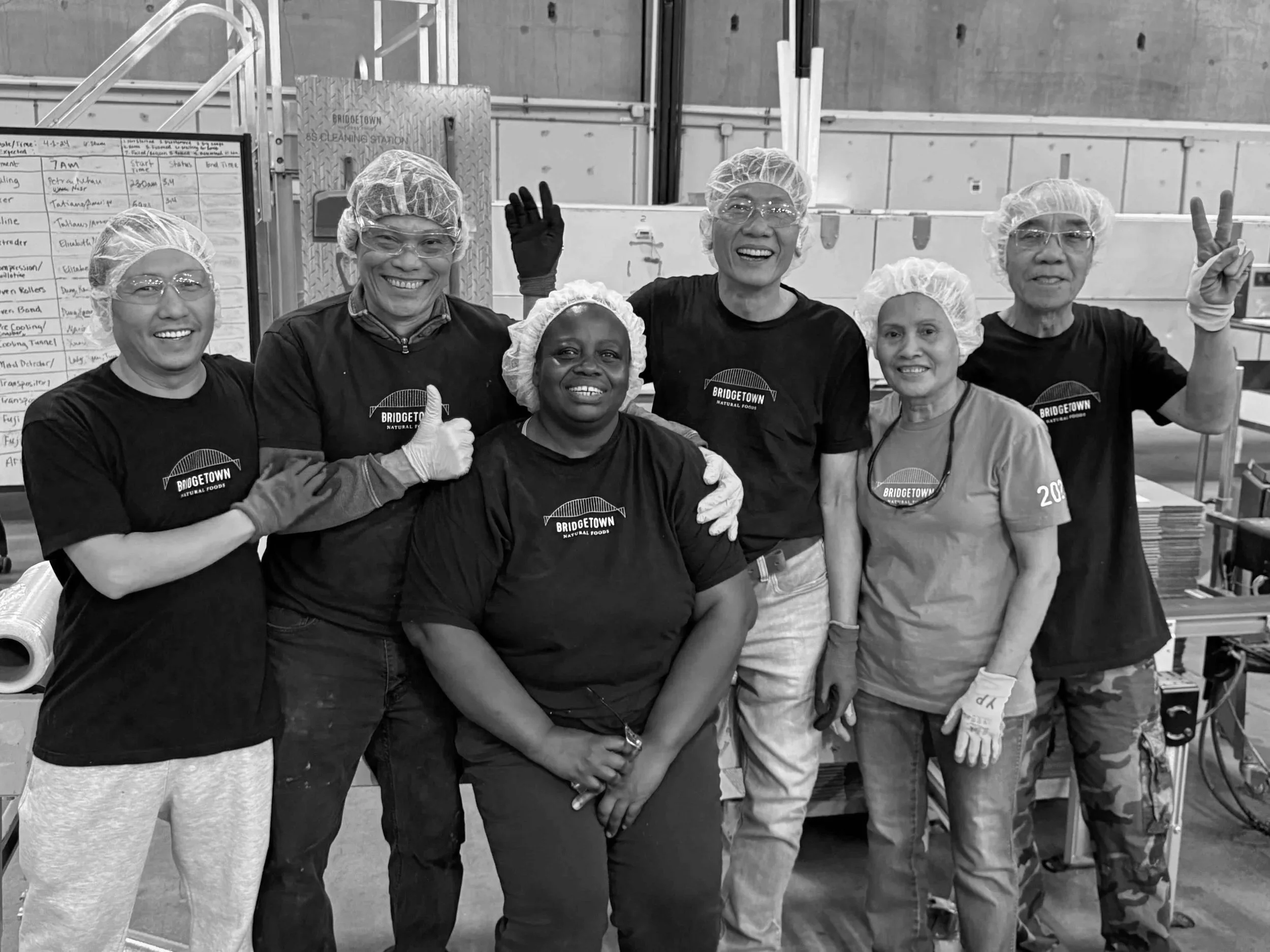 Group of seven people in a commercial kitchen wearing hairnets, gloves, and black T-shirts with 'Bridgetown Natural Foods' logo, smiling and posing for the photo.