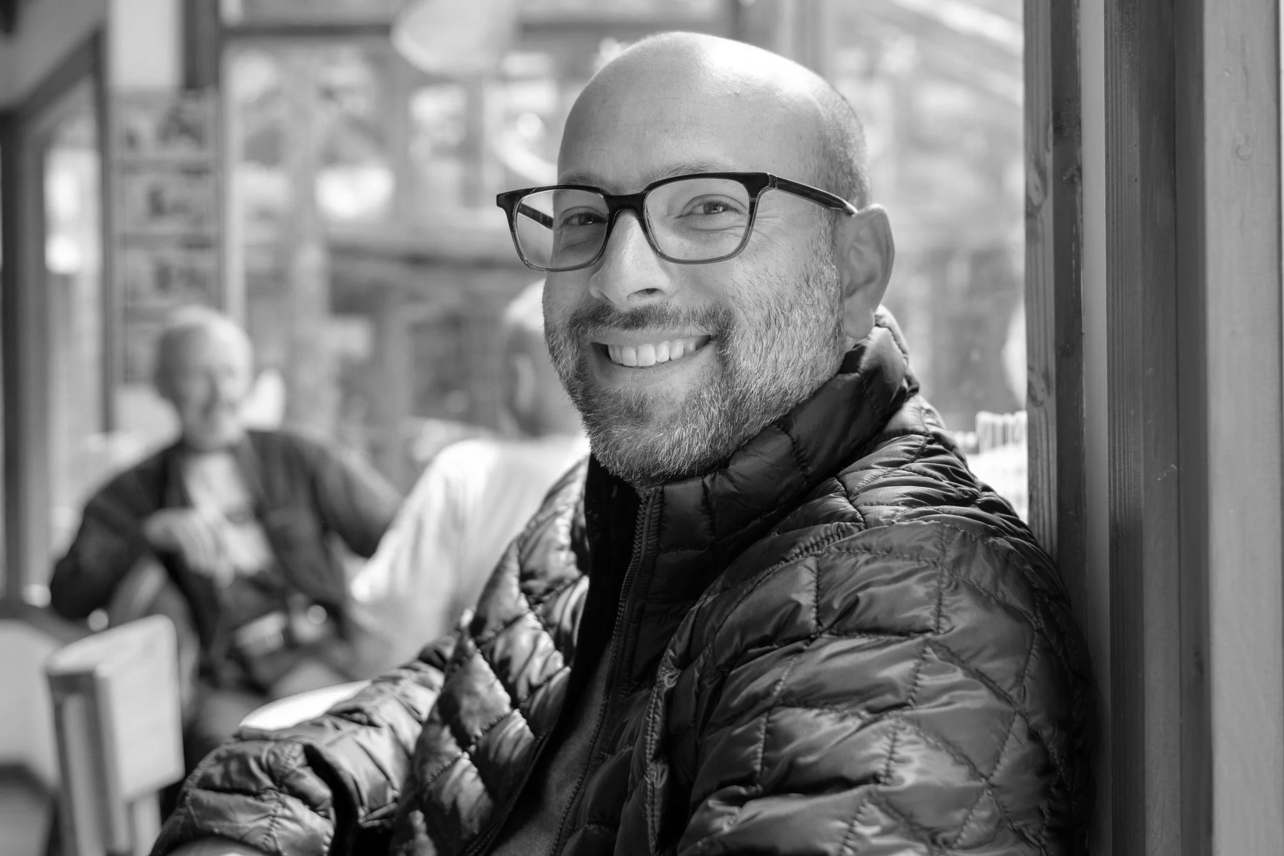 A smiling man with glasses and a beard, wearing a puffy jacket, sitting in a wooden structure with blurred people in the background.