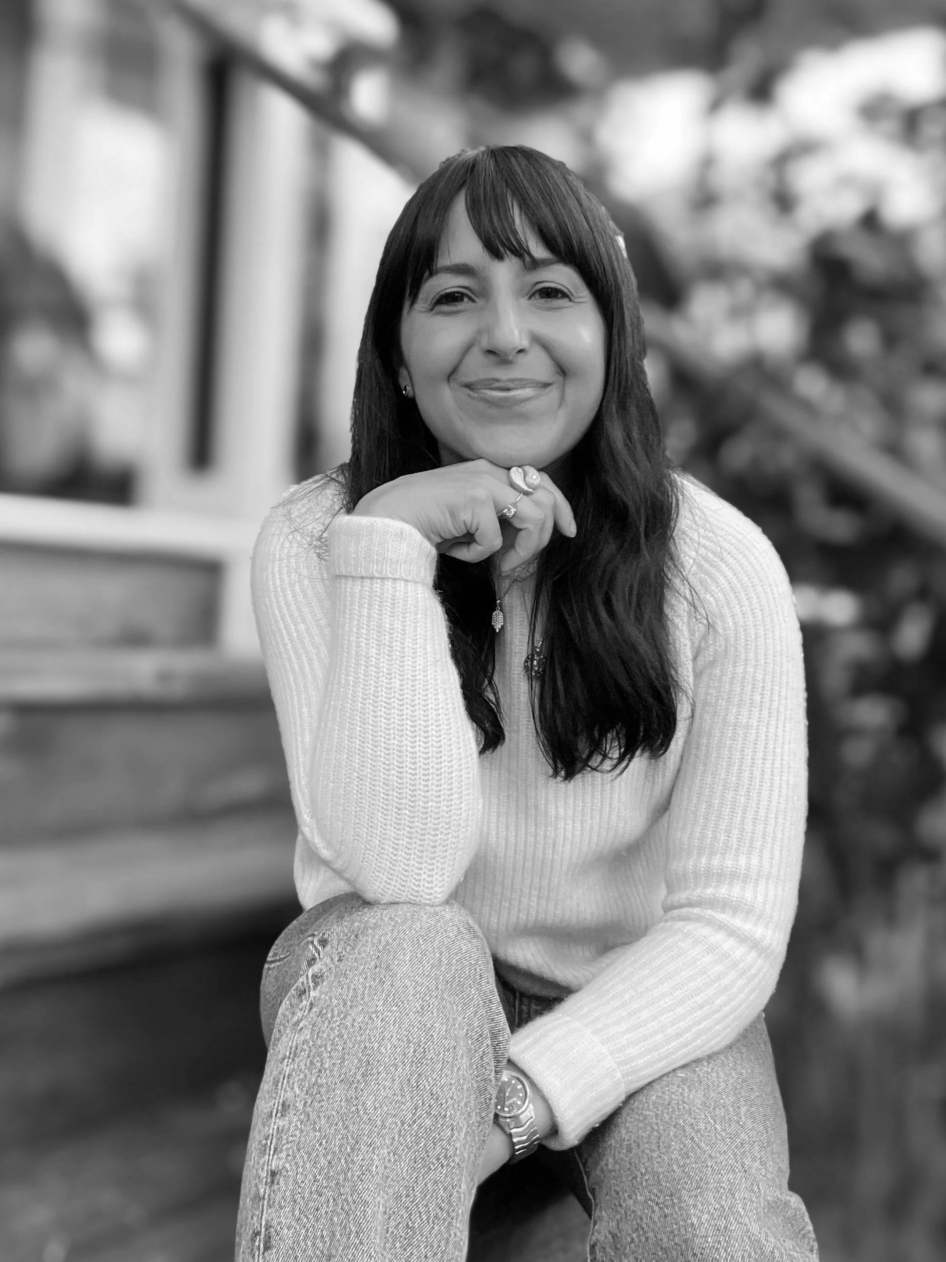 Black and white photo of a smiling woman sitting outside with one hand resting on her chin, wearing a light-colored sweater, jeans, a wristwatch, and rings, with a blurred background of foliage and stairs.
