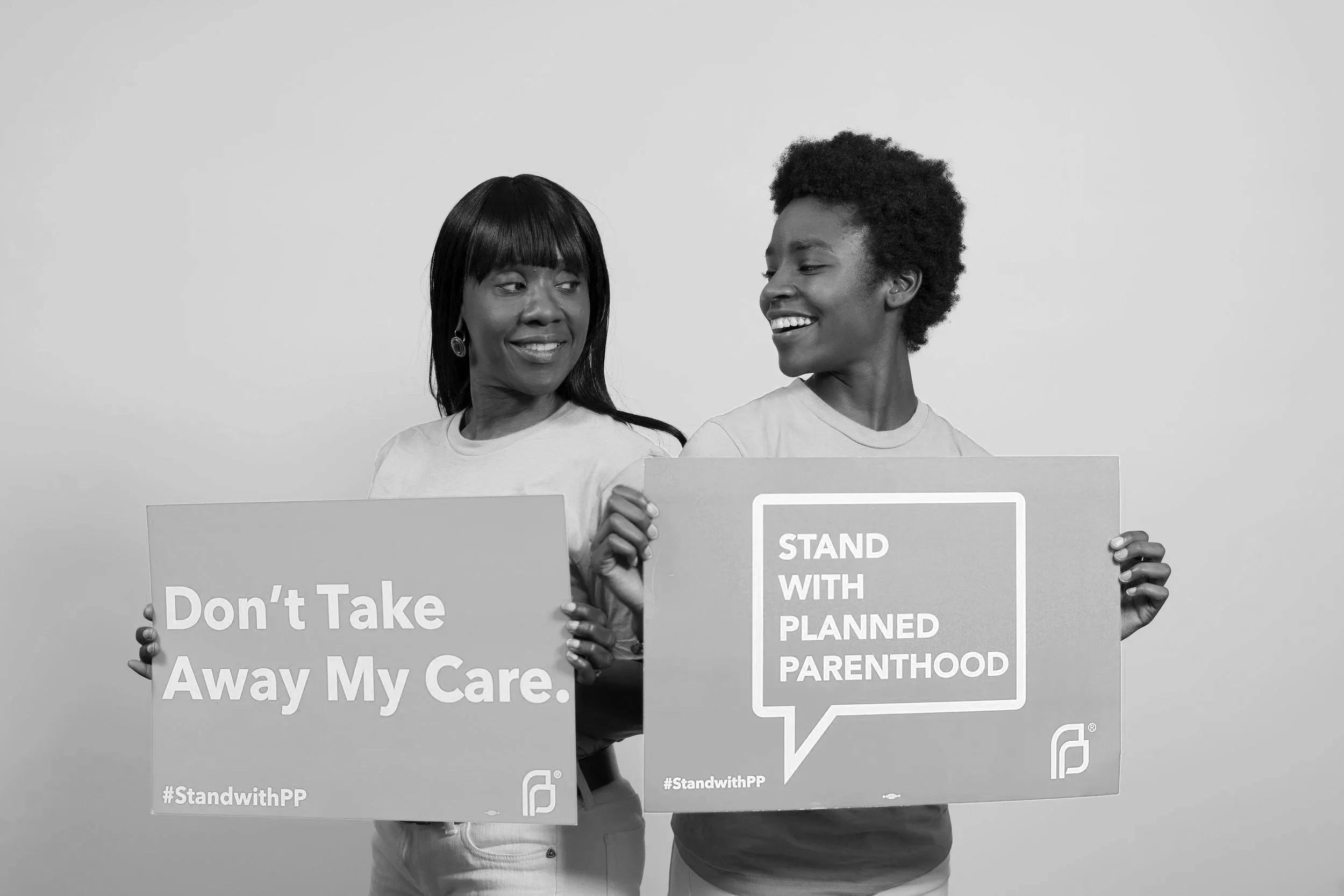 Two women holding protest signs, one sign reads 'Don't Take Away My Care,' the other reads 'STAND WITH PLANNED PARENTHOOD,' smiling at each other.