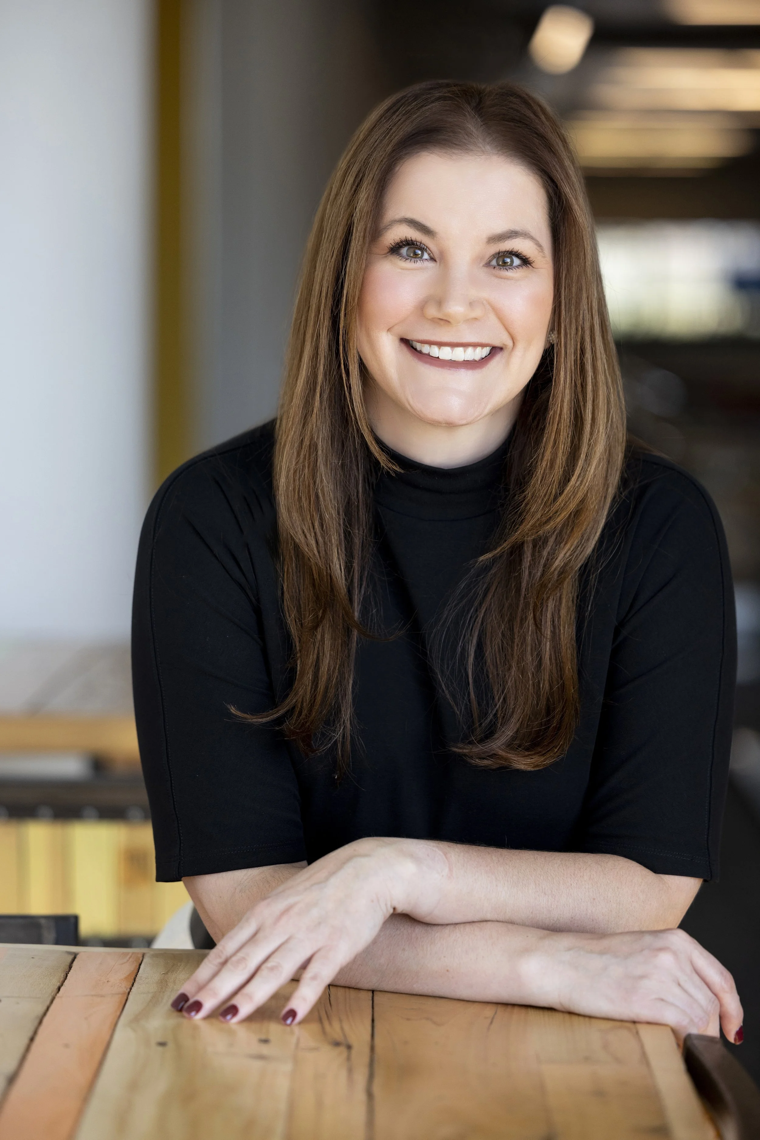 A woman with long brown hair and a black turtleneck sweater, smiling and sitting at a table in a casual indoor setting.