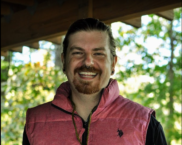 A man with long hair, a beard, and a mustache smiling outdoors under a wooden structure with green trees in the background, wearing a pink vest over a dark shirt.
