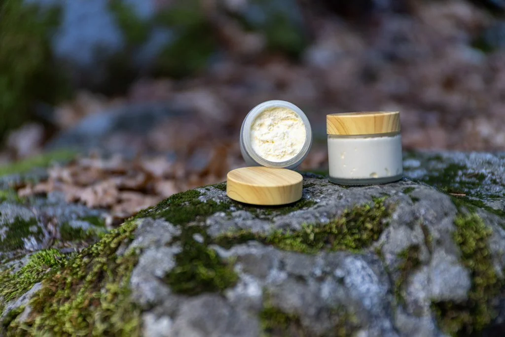 Open jar of cream or lotion with a wooden lid next to another closed jar with a similar lid, placed on a moss-covered rock in a forest setting.