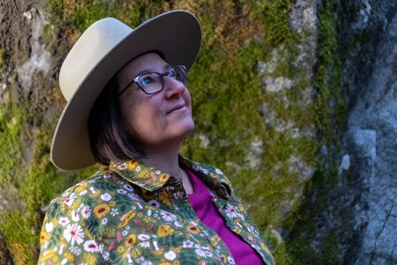 A woman wearing glasses, a wide-brimmed hat, and a floral jacket standing outdoors near moss-covered rocks.