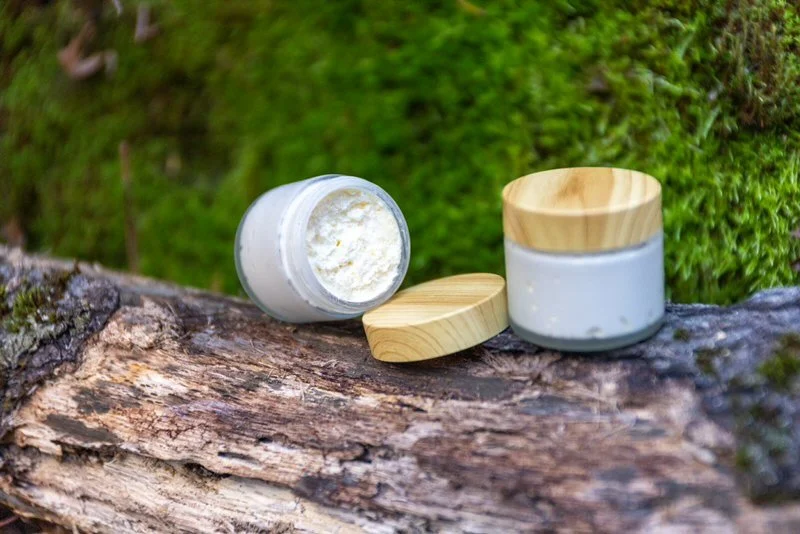 Open container of white cream or lotion with wooden lid, placed on a mossy log outdoors.