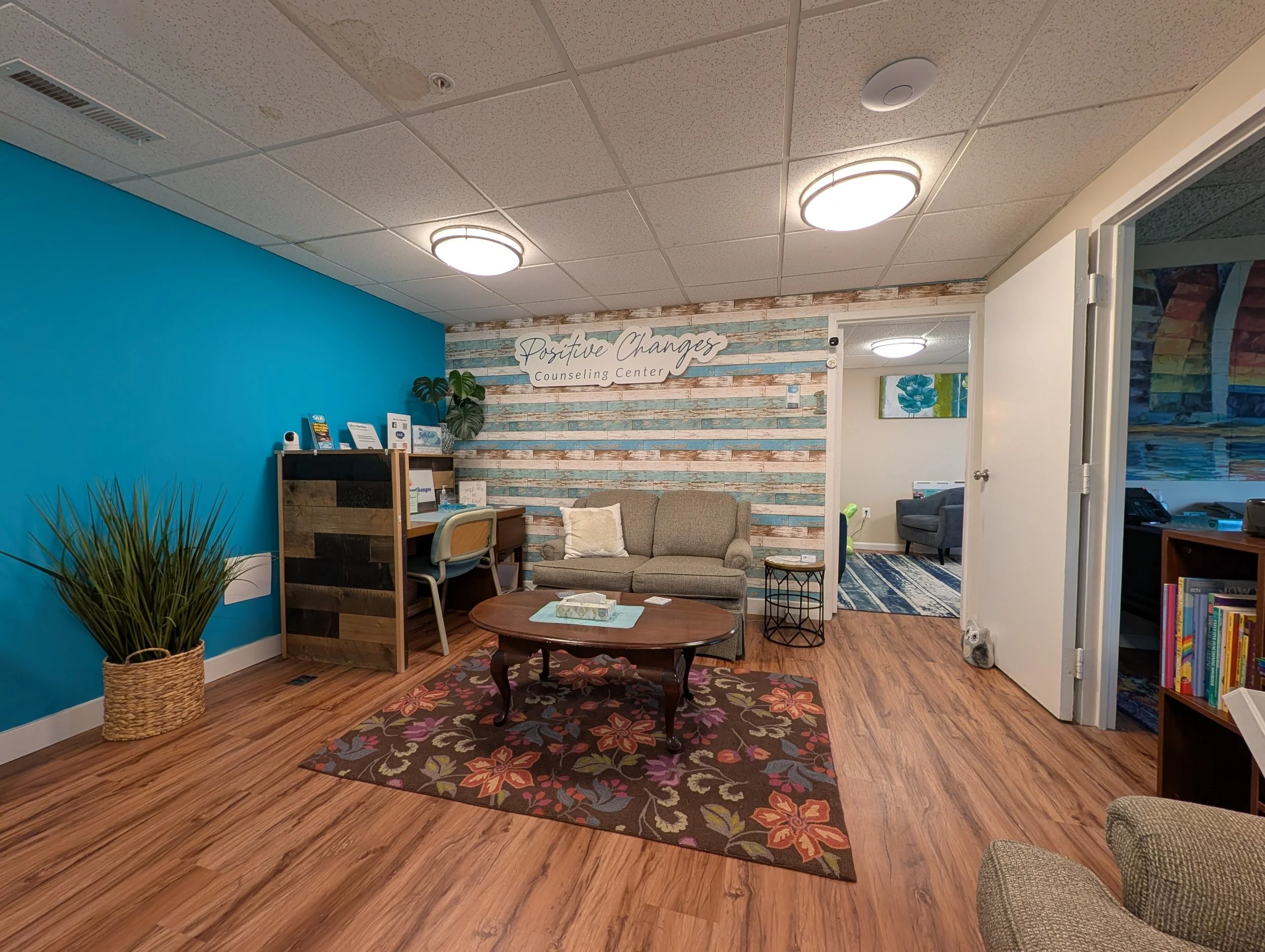 Waiting room of a therapy office with a sign reading 'Positive Changes Counseling Center,' a teal accent wall, a wooden floor, a small floral area rug, a wooden coffee table, a sofa, a bookshelf with books, a potted plant, and artwork on the walls.