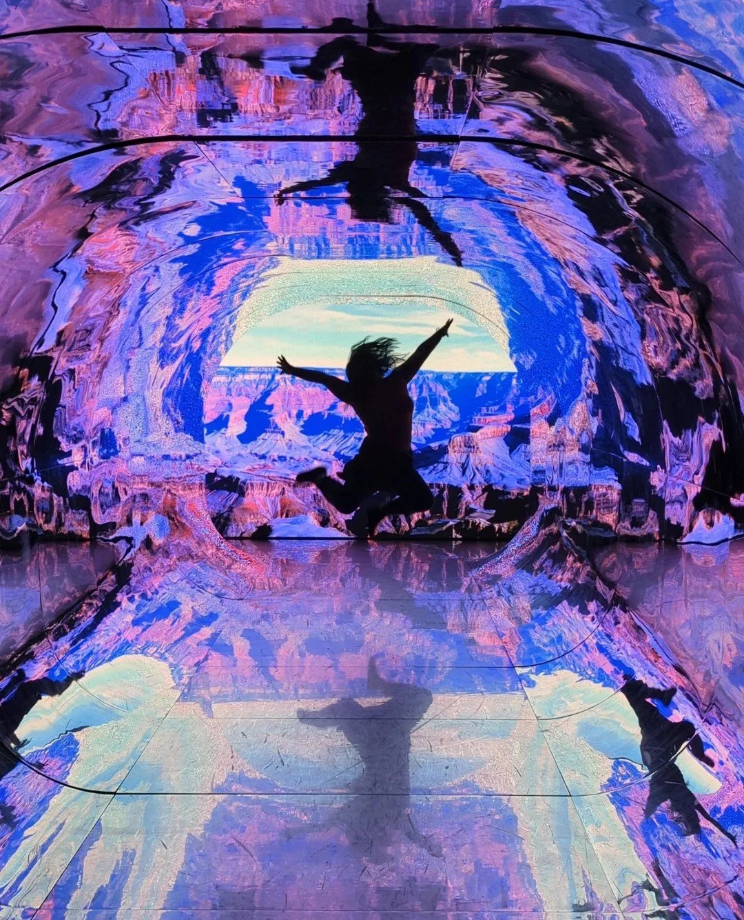 A person jumping inside a colorful, illuminated tunnel with abstract patterns, creating a silhouette with arms and legs spread out and a reflection on the tunnel's shiny surface.