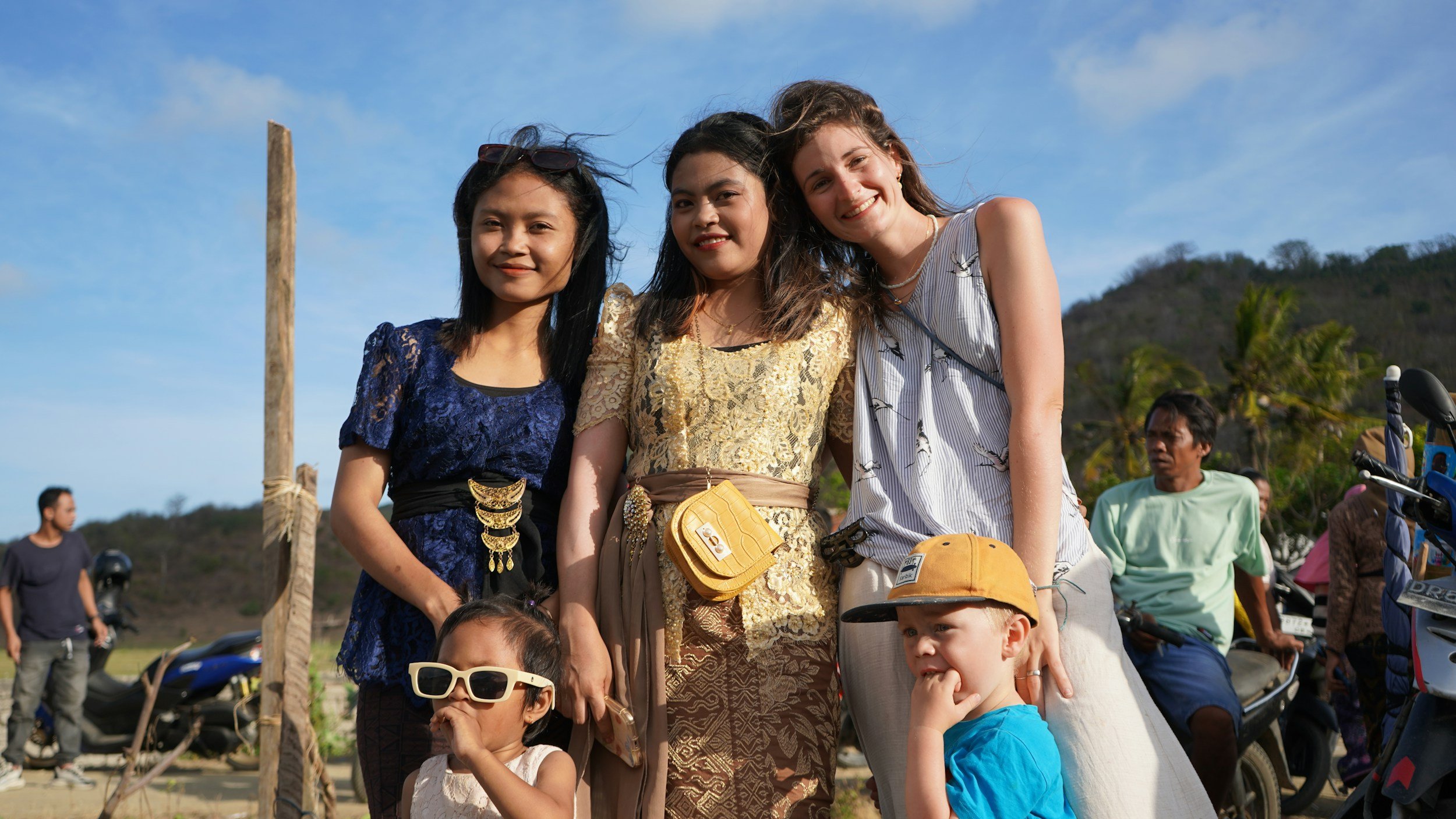 Group of smiling women and children outdoors on a sunny day with motorcycles and a hilly landscape in the background.