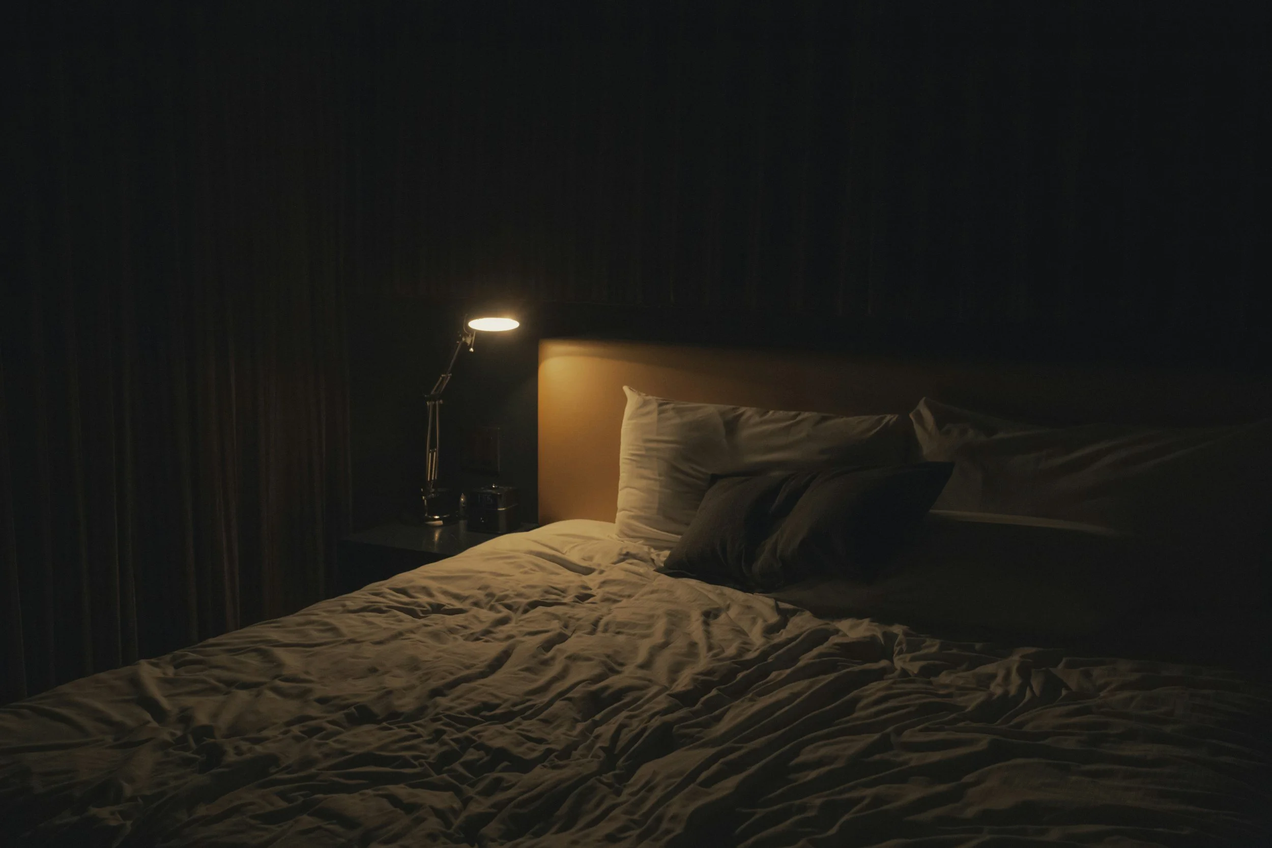 A dimly lit bedroom with a bed, a bedside table, and a lamp turned on.