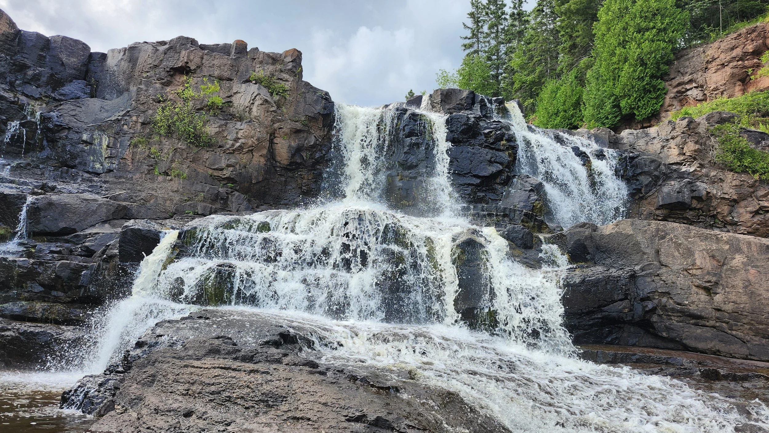 A scenic view of a rocky waterfall with water cascading down over dark rocks, surrounded by green trees and bushes under a partly cloudy sky.