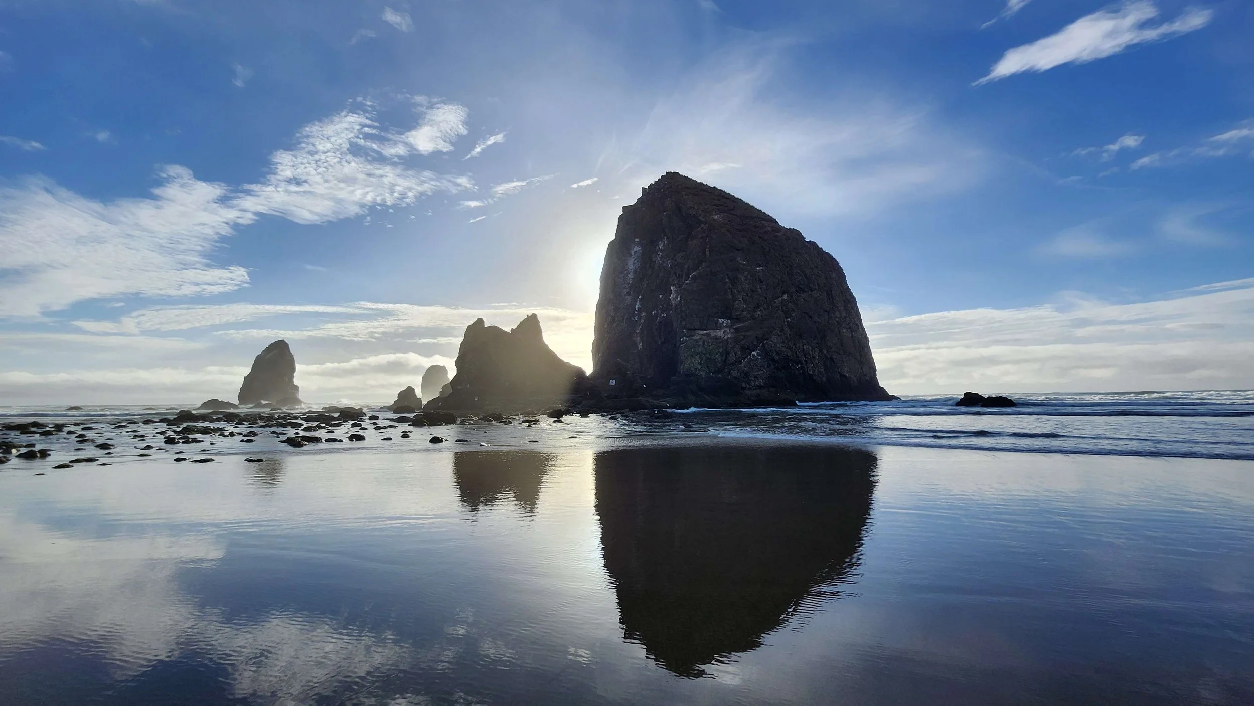Sea stacks on the beach with reflection in the wet sand, partly cloudy sky, and sun near the horizon.