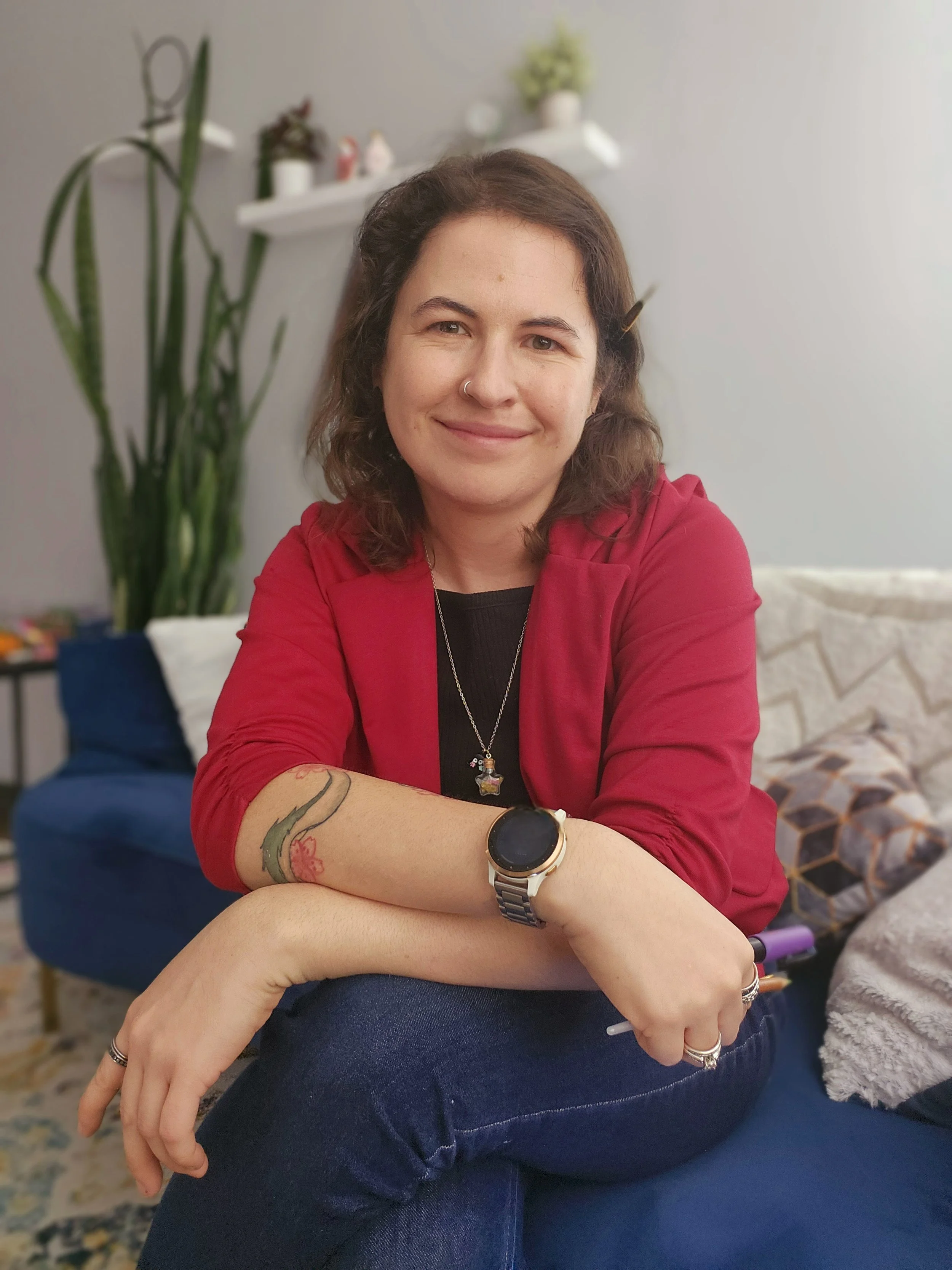 A woman with shoulder-length brown hair, a nose ring, and tattoos on her left forearm, wearing a red jacket, a black shirt, a watch, and jewelry, sitting on a couch in a cozy living room.