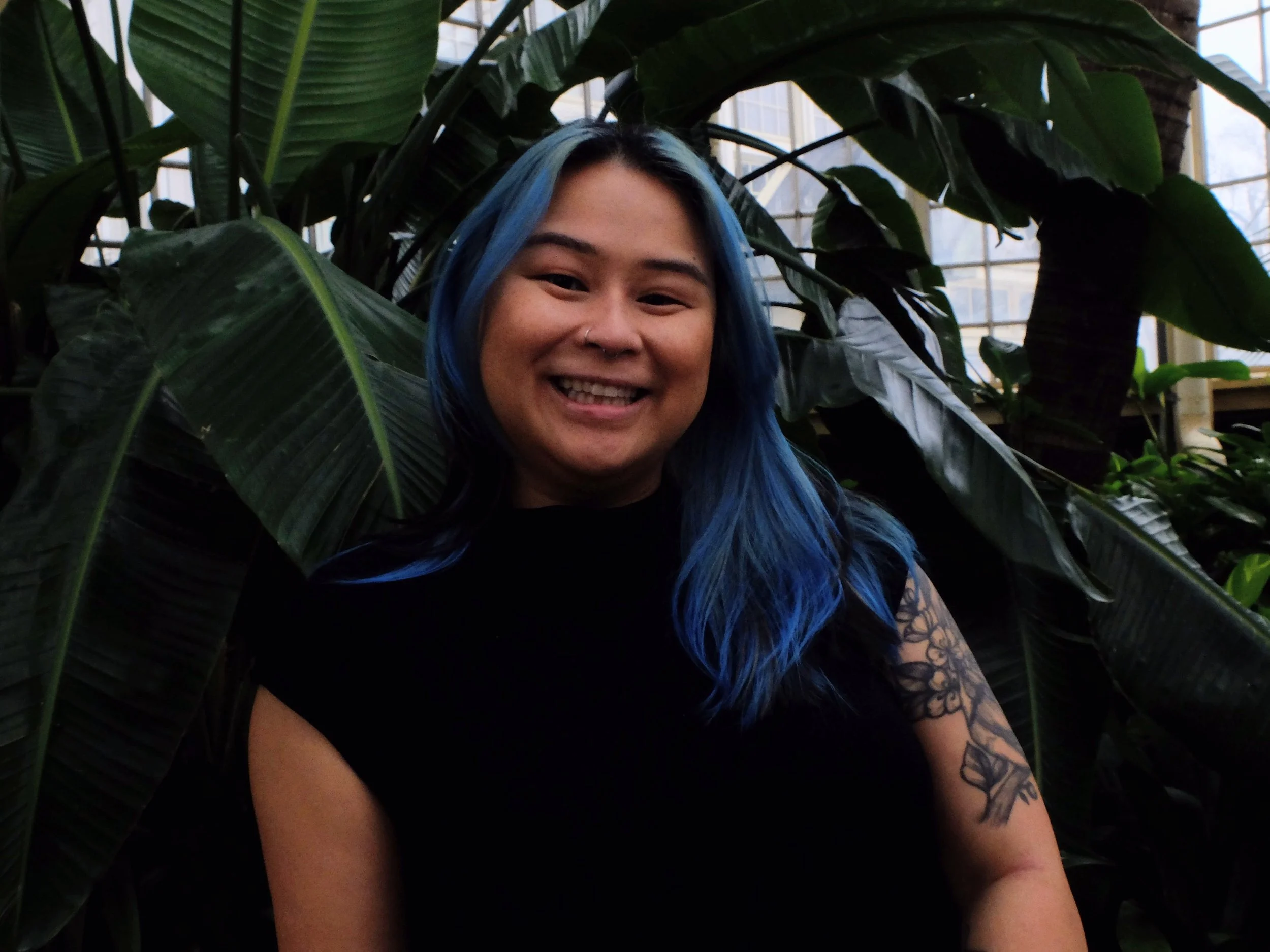 A young woman with shoulder-length hair dyed bright blue and black. She is smiling slightly, wearing a black T-shirt with a graphic of two raisins on the front. She has a small nose ring and a natural complexion. The background features a blurred indoor setting with some furniture and a large fish tank.