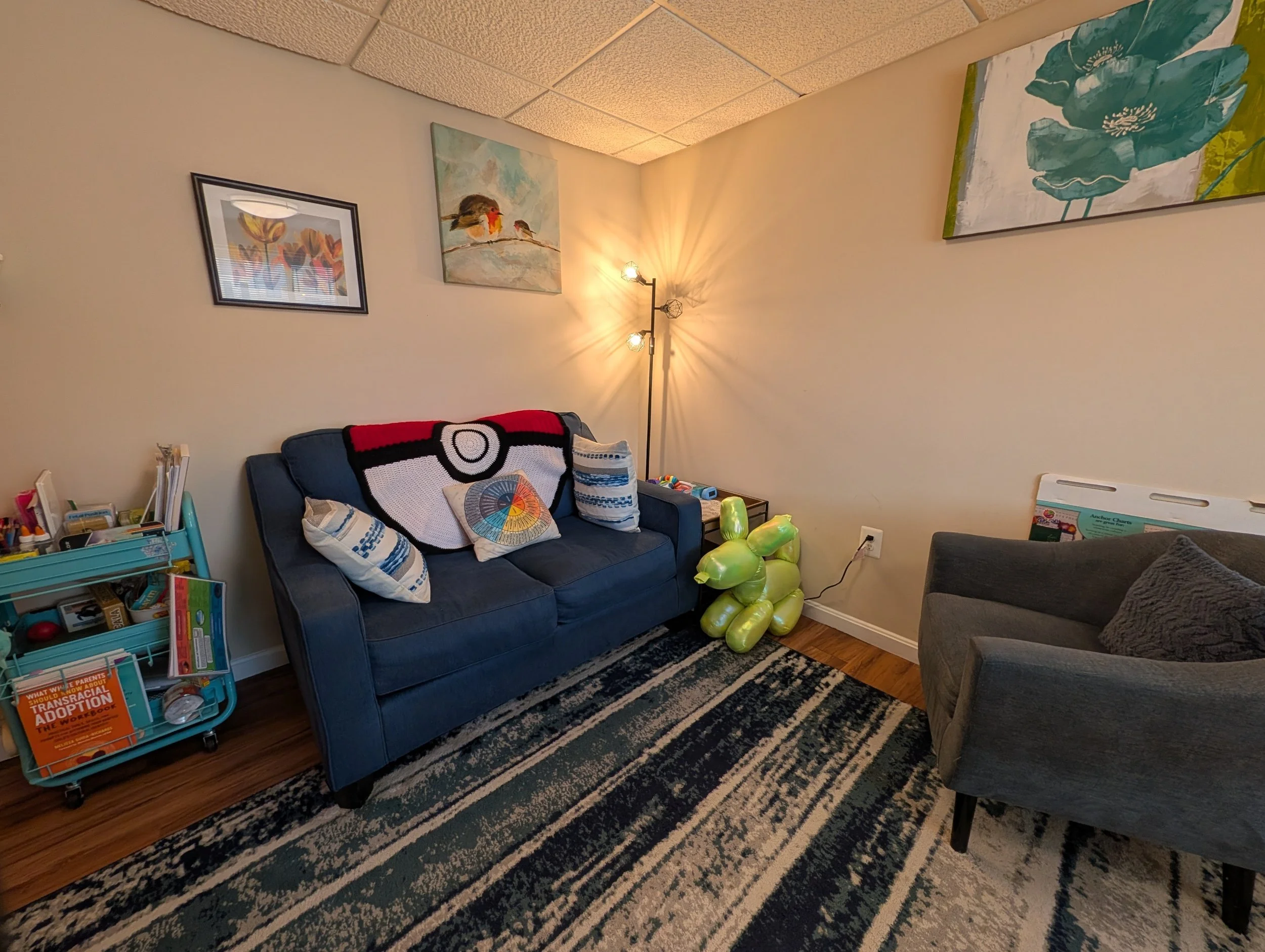 A cozy therapy office corner featuring wall art with birds and flowers, a navy blue sofa with patterned pillows, a floor lamp with multiple bulbs, a rug with stripes, a navy/grey armchair, and a small teal utility cart with books and games.