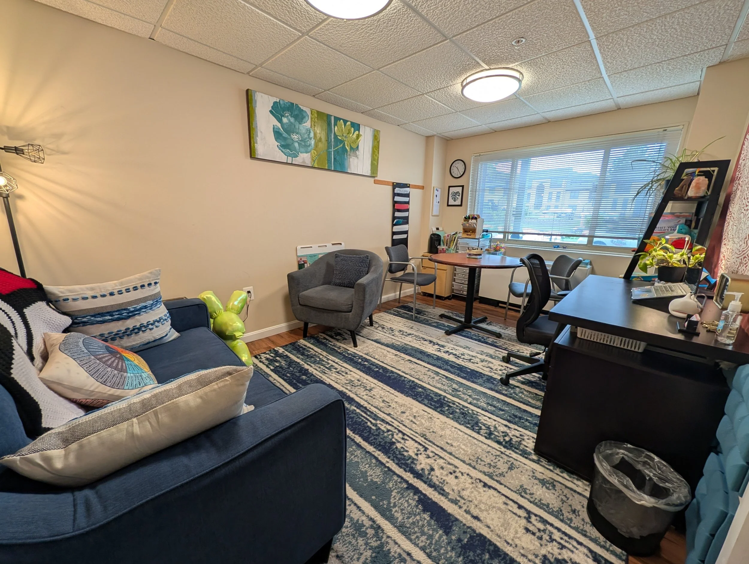 Office with a blue sofa and pillows, a rug, a window with blinds, a round table with chairs, shelves, wall art, and various office supplies.
