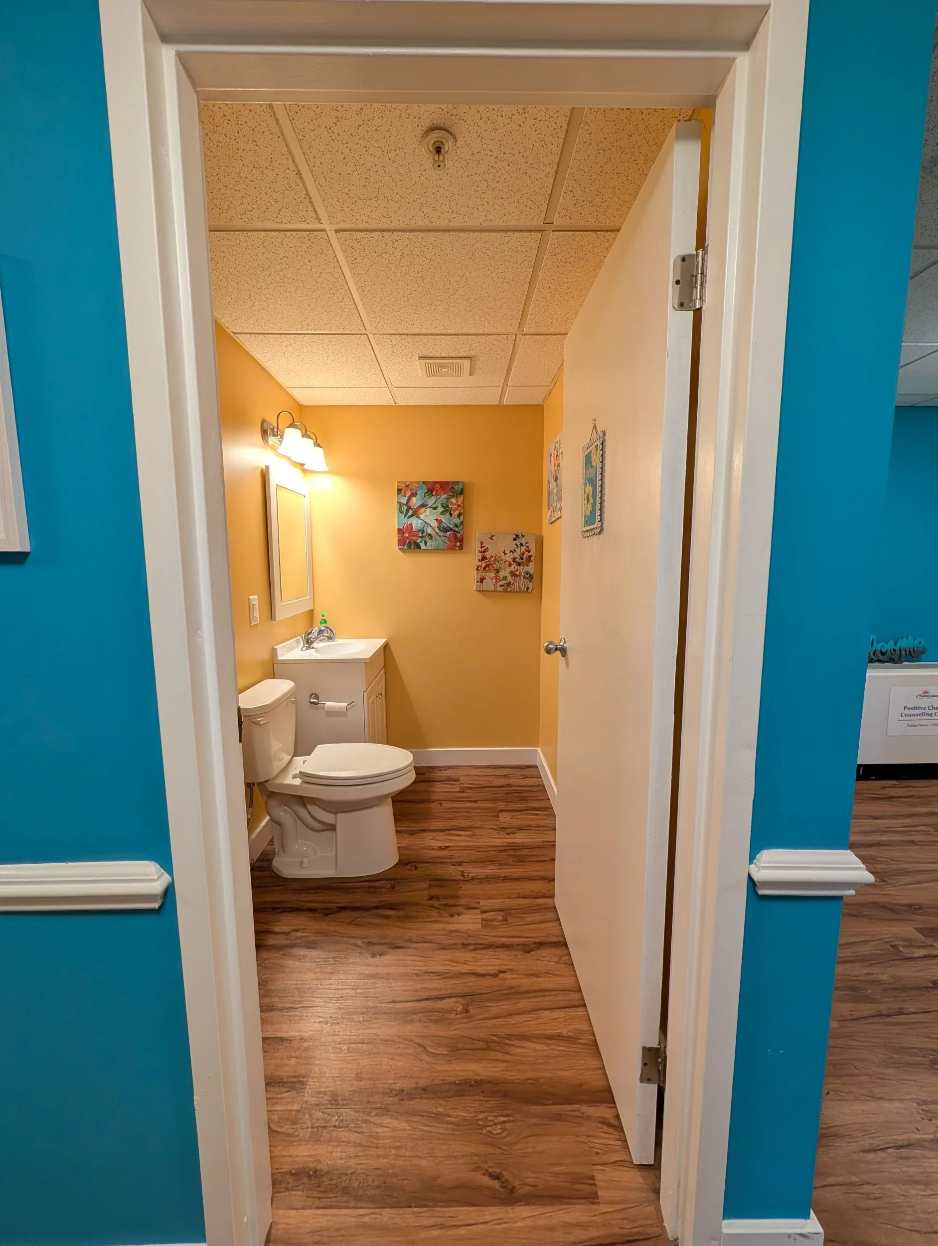 View of a bathroom with yellow walls, ceiling tiles, light fixture, framed artwork, white toilet, and bathroom sink, through an open door with blue walls surrounding the doorway.