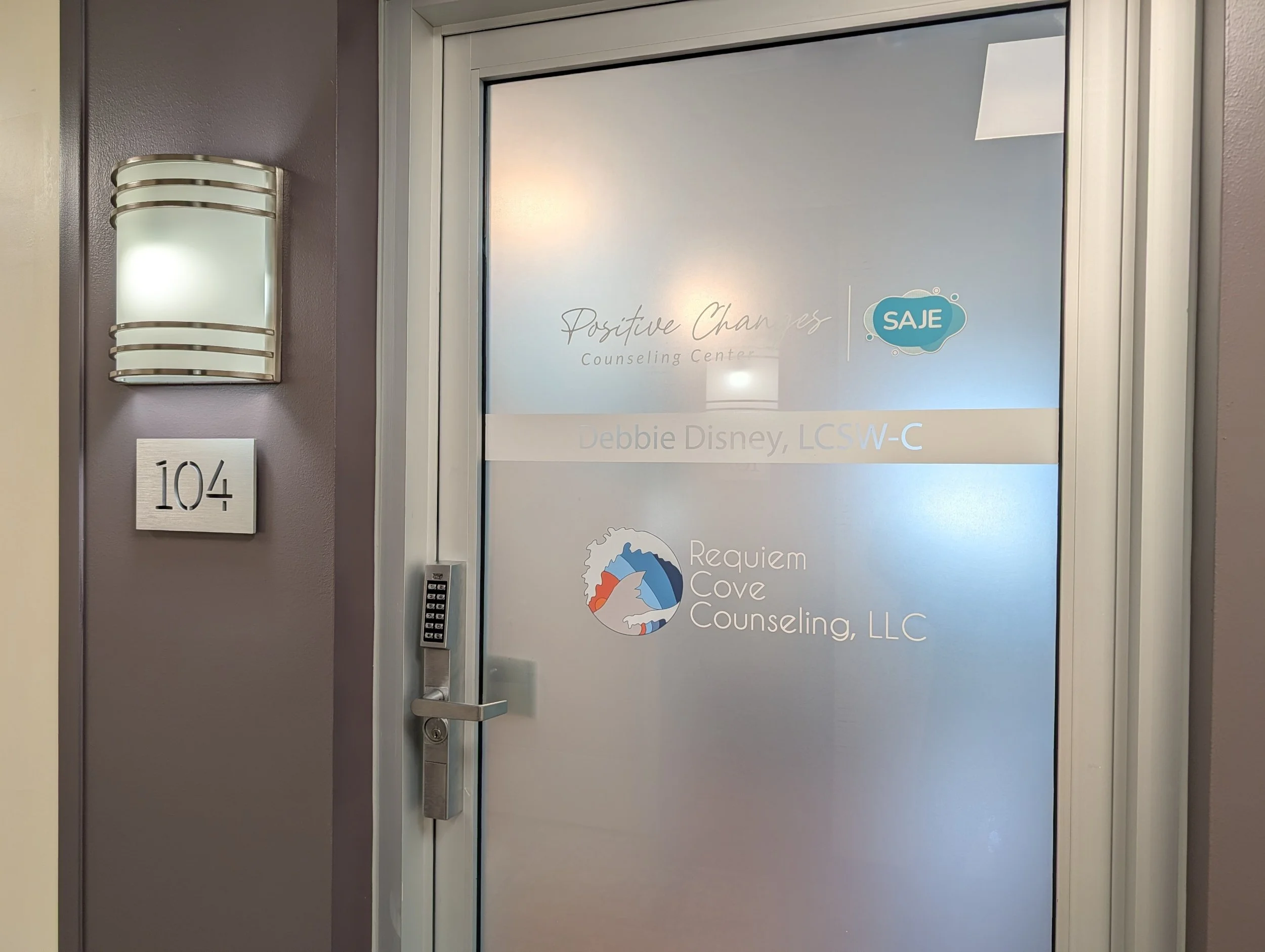 An office door with frosted glass, labeled "Positive Change Counseling Center | SAJE" and "Requiem Cove Counseling". The door handle is on the left and has a keypad. To the left, there is a wall-mounted light fixture and a sign that reads "104."