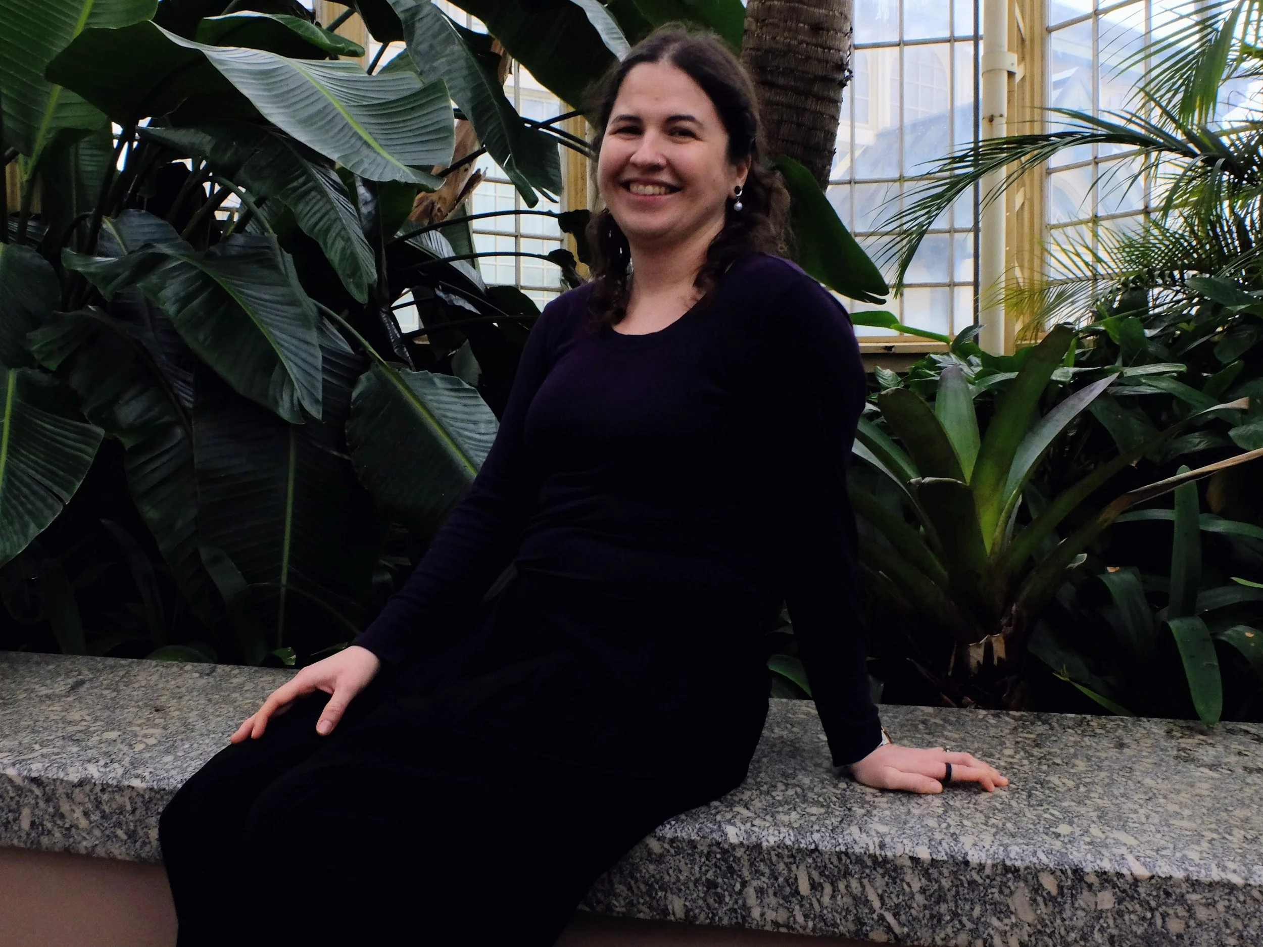 A woman with shoulder-length brown hair, a nose ring, and tattoos on her left forearm, wearing a red jacket, a black shirt, a watch, and jewelry, sitting on a couch in a cozy living room.